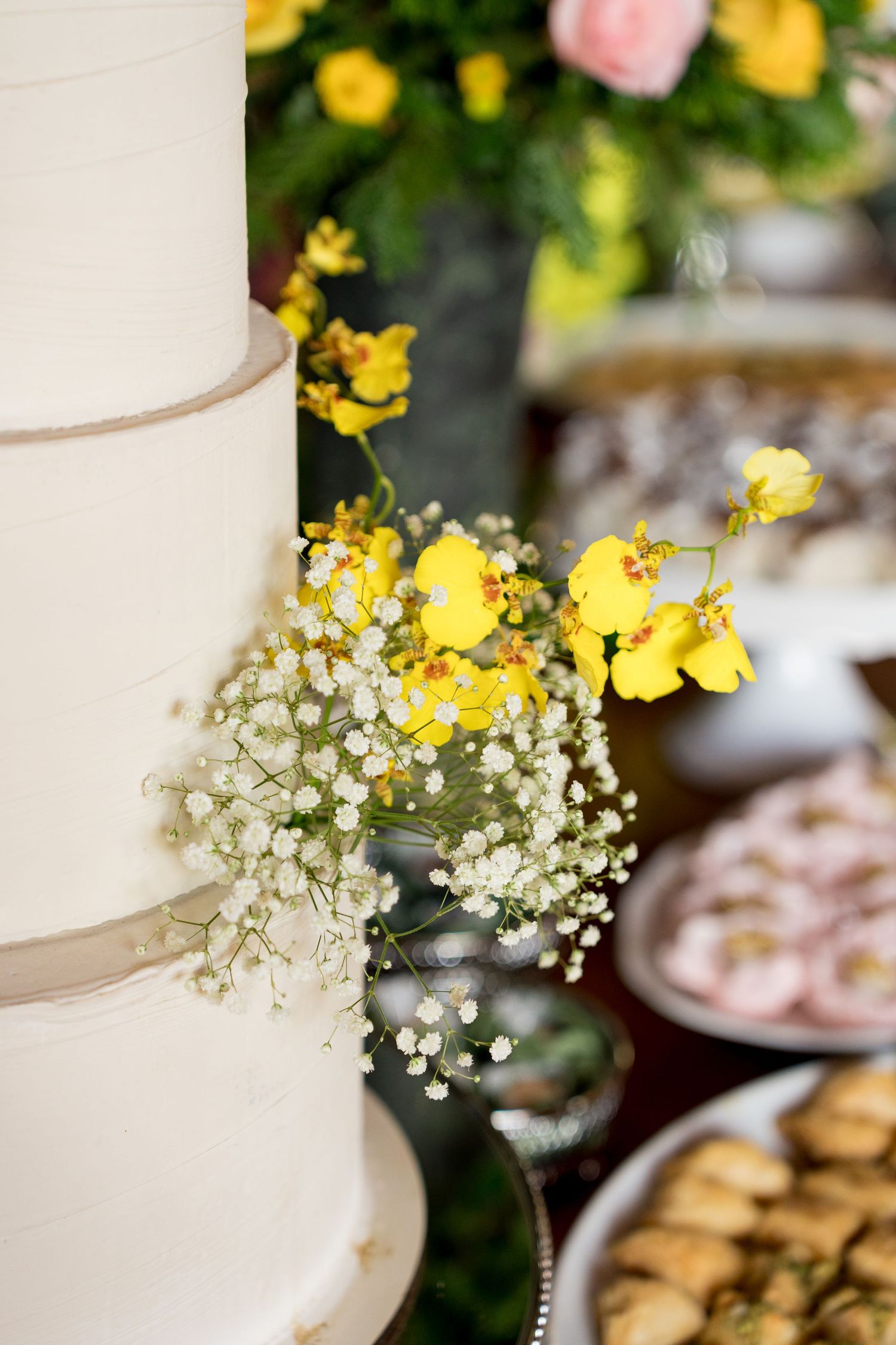 White tiered wedding cake adorned with yellow orchids and baby's breath flowers, with dessert table in background.