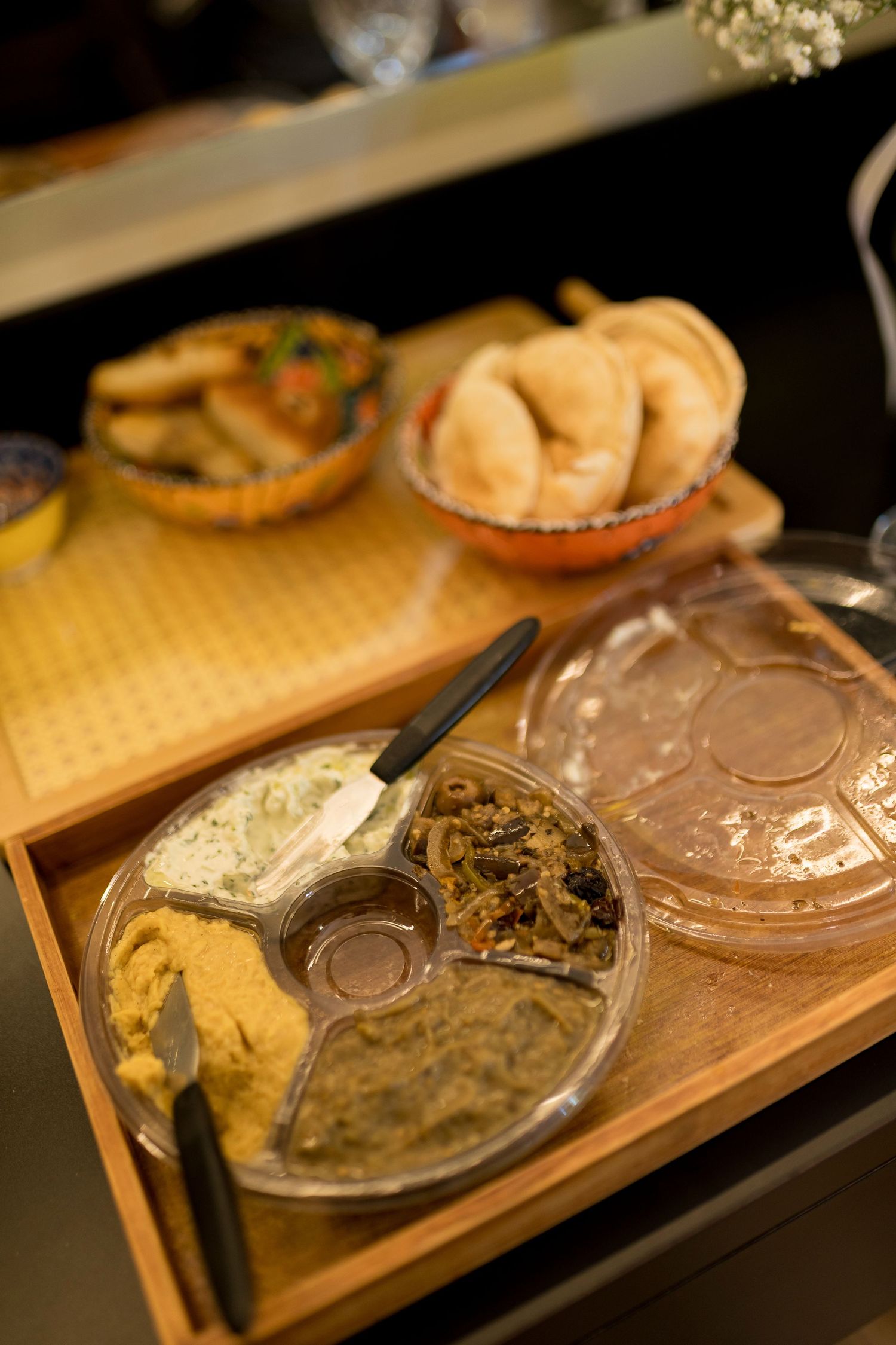 A wooden tray with a divided plastic platter of hummus, greens, and dips, alongside bread and appetizers in the background.