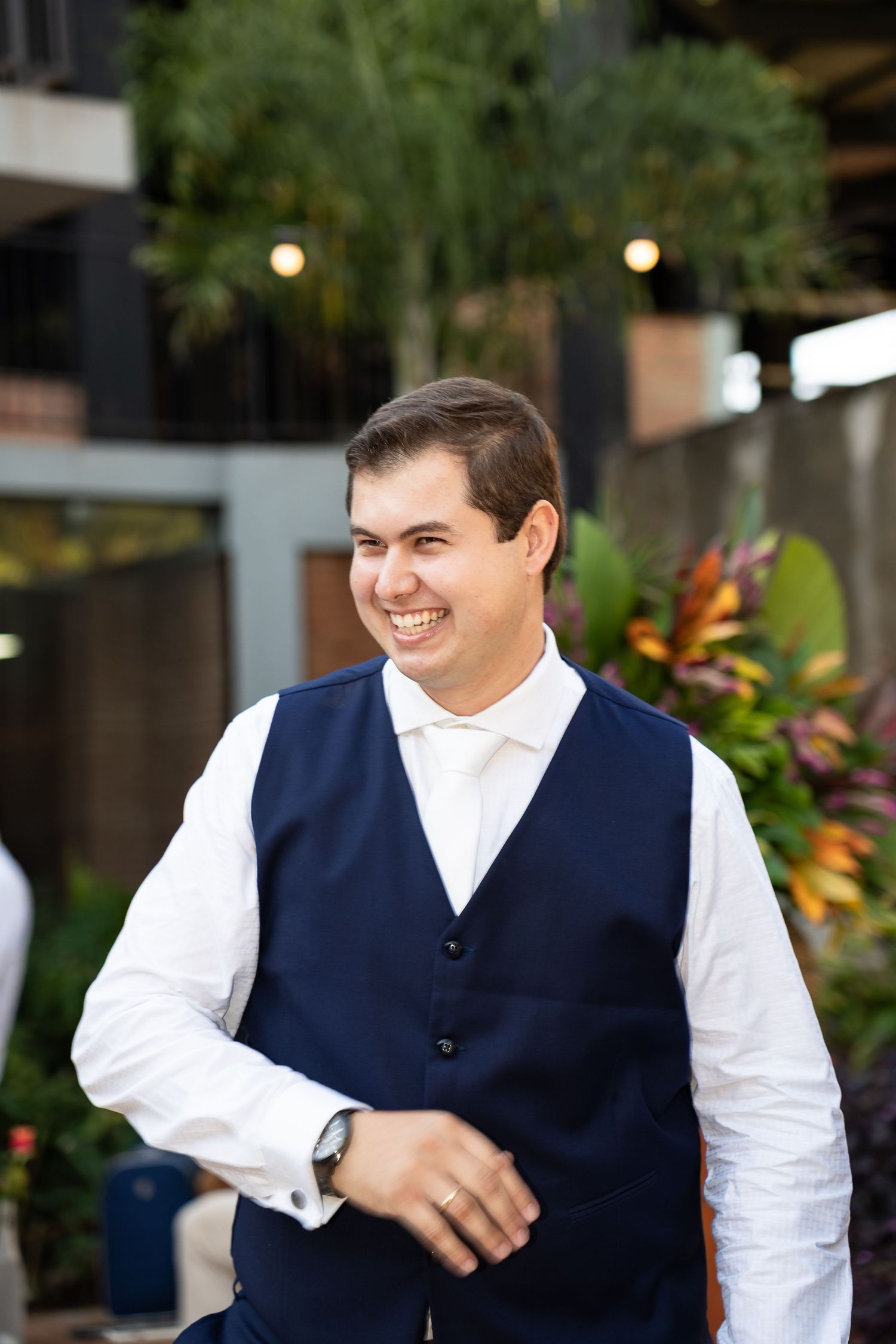 Smiling groom in navy blue vest and white shirt with tie, standing outdoors surrounded by tropical greenery.