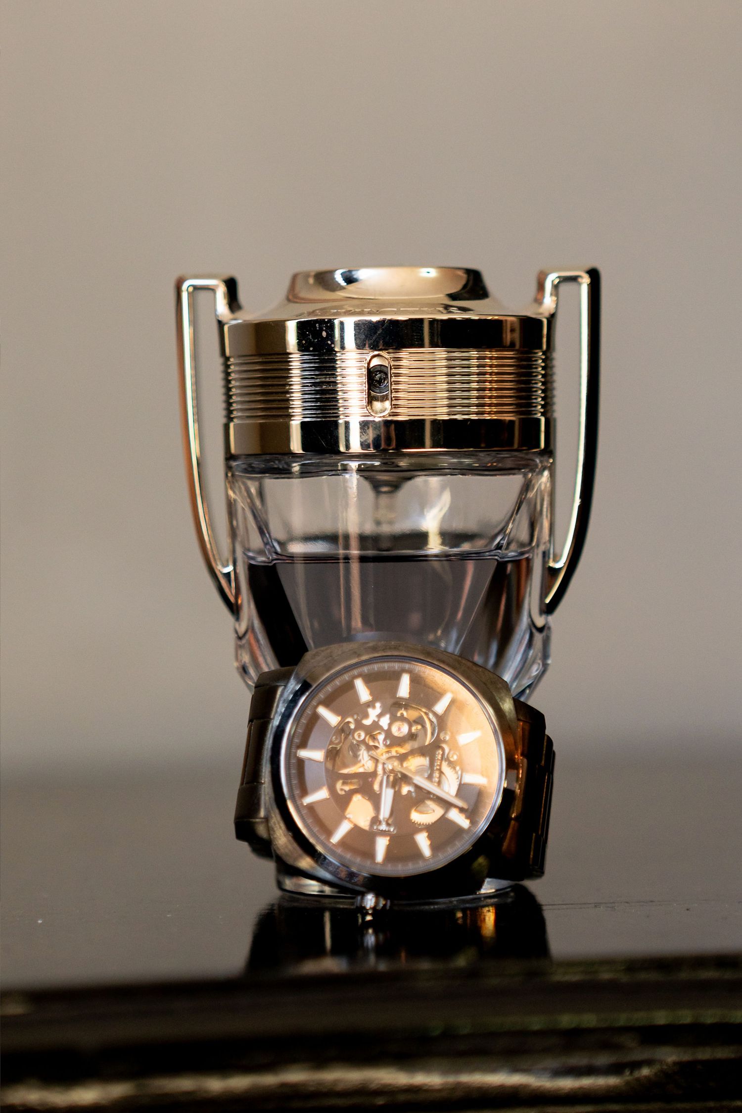 A luxury trophy-shaped perfume bottle with a skeleton watch placed in front on a reflective surface.