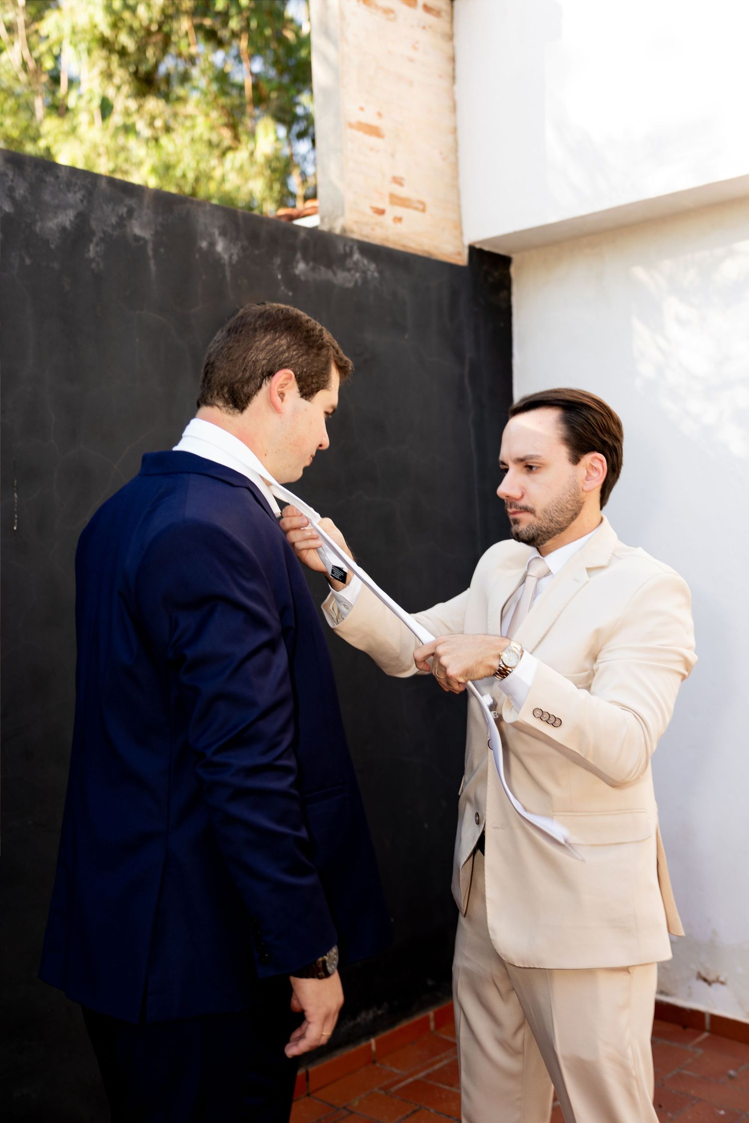 Man in beige suit helping groom in navy suit adjust his tie outdoors before wedding ceremony.