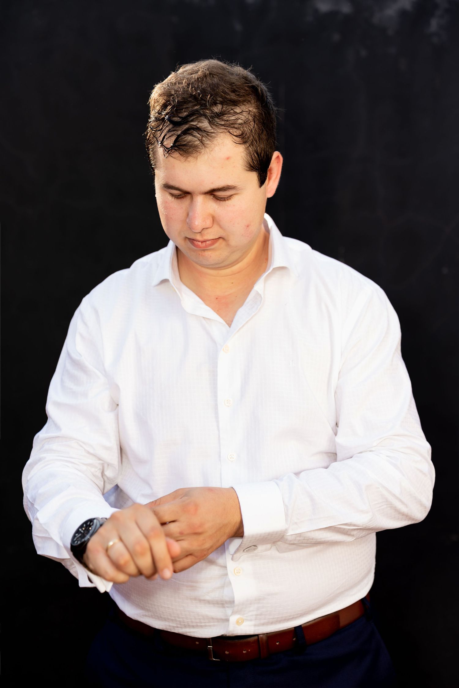 Man in white dress shirt adjusting his cufflinks against a dark background.