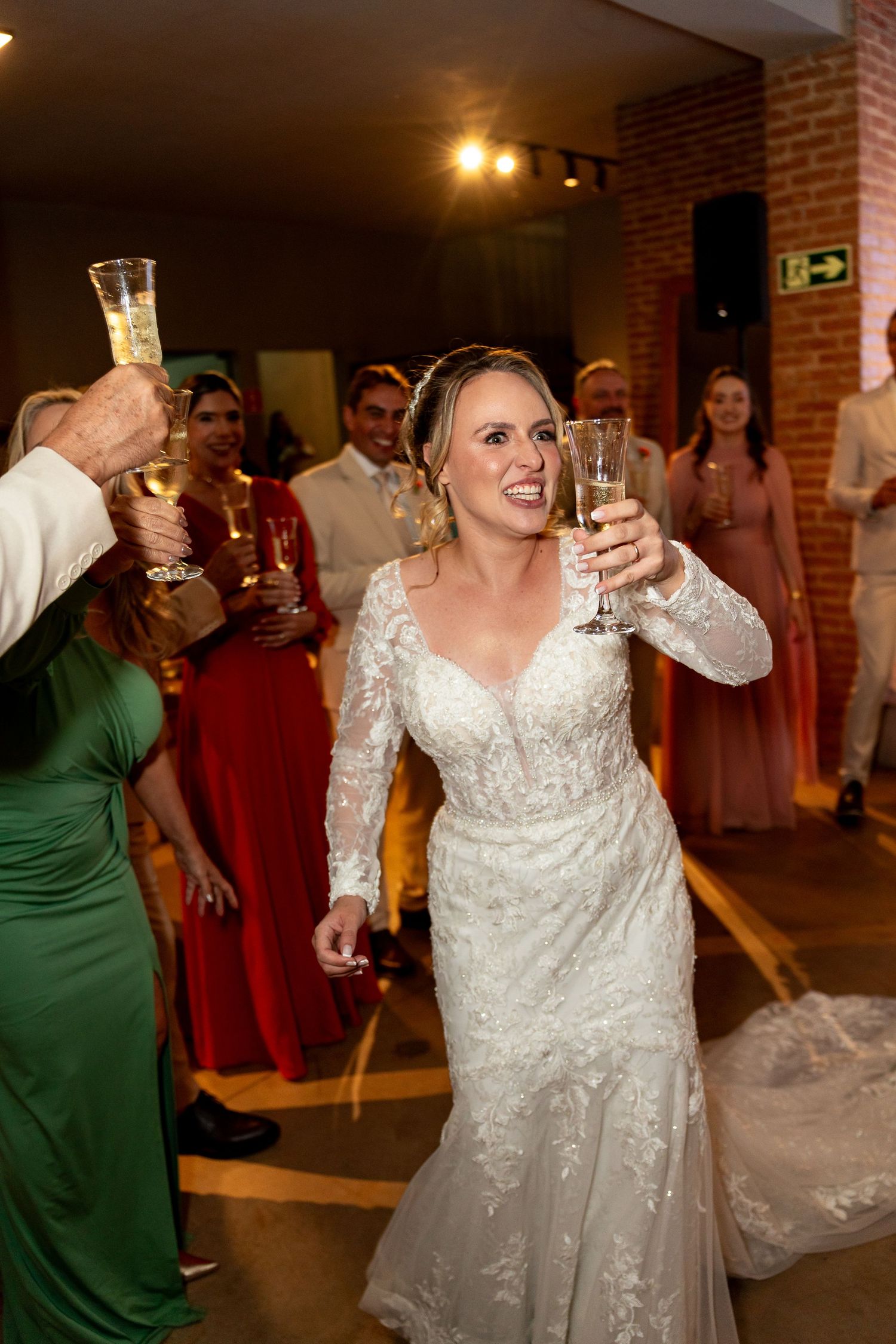 Bride in lace gown raises champagne glass during wedding reception toast surrounded by guests in formal attire.