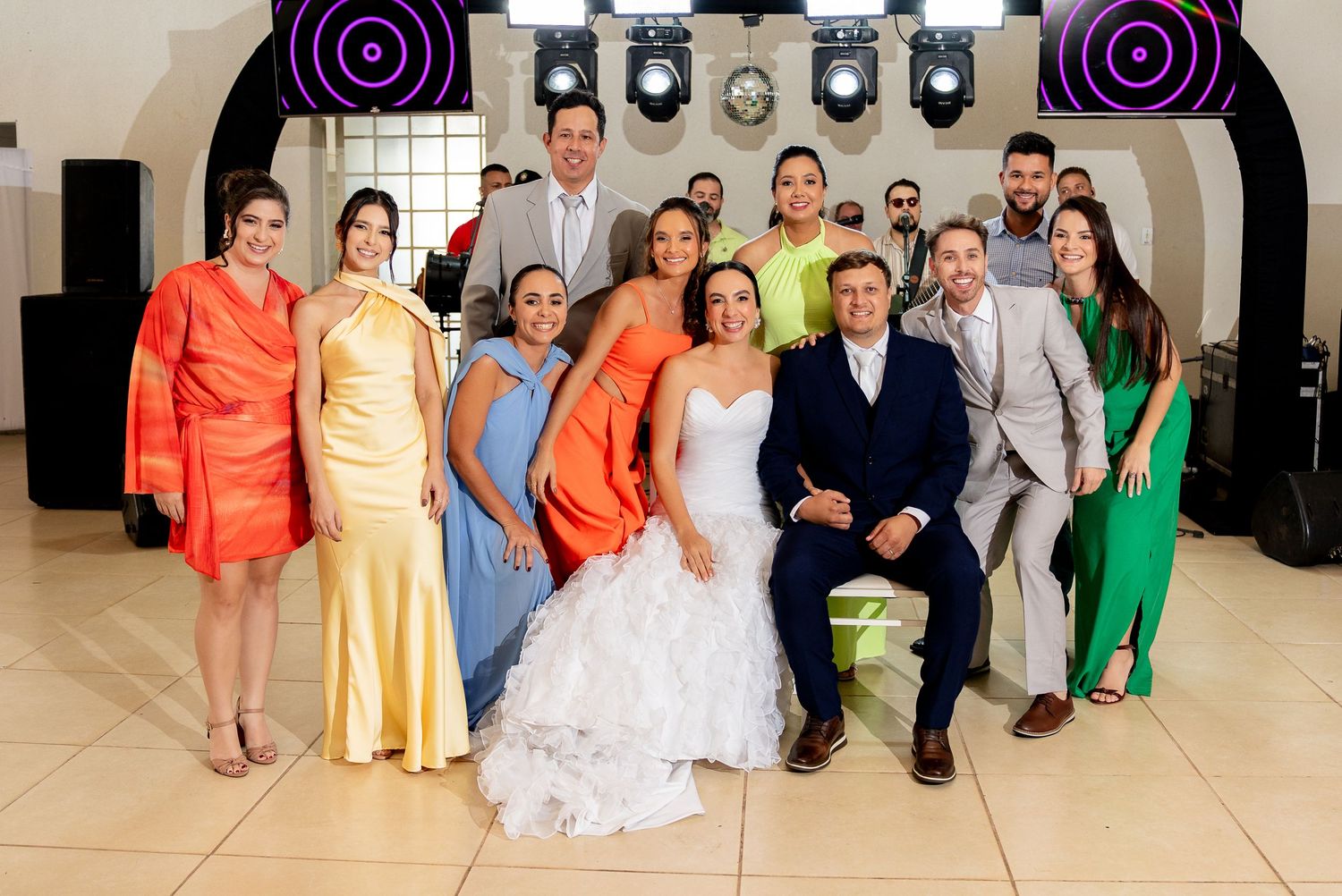Wedding party group photo with bride in white gown, groom, and guests in colorful formal attire at a reception venue.