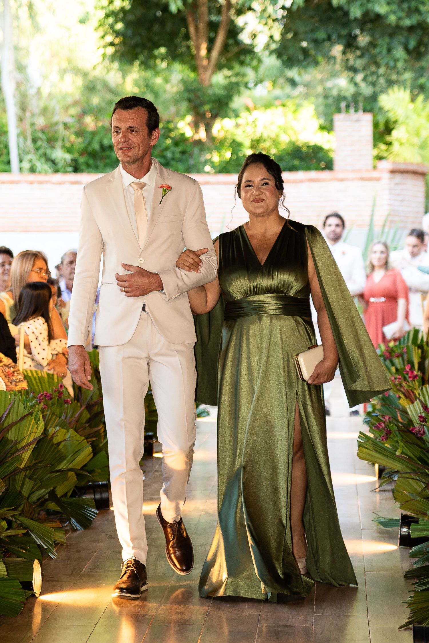 Groom in white suit walks with woman in elegant green cape gown at outdoor wedding ceremony.