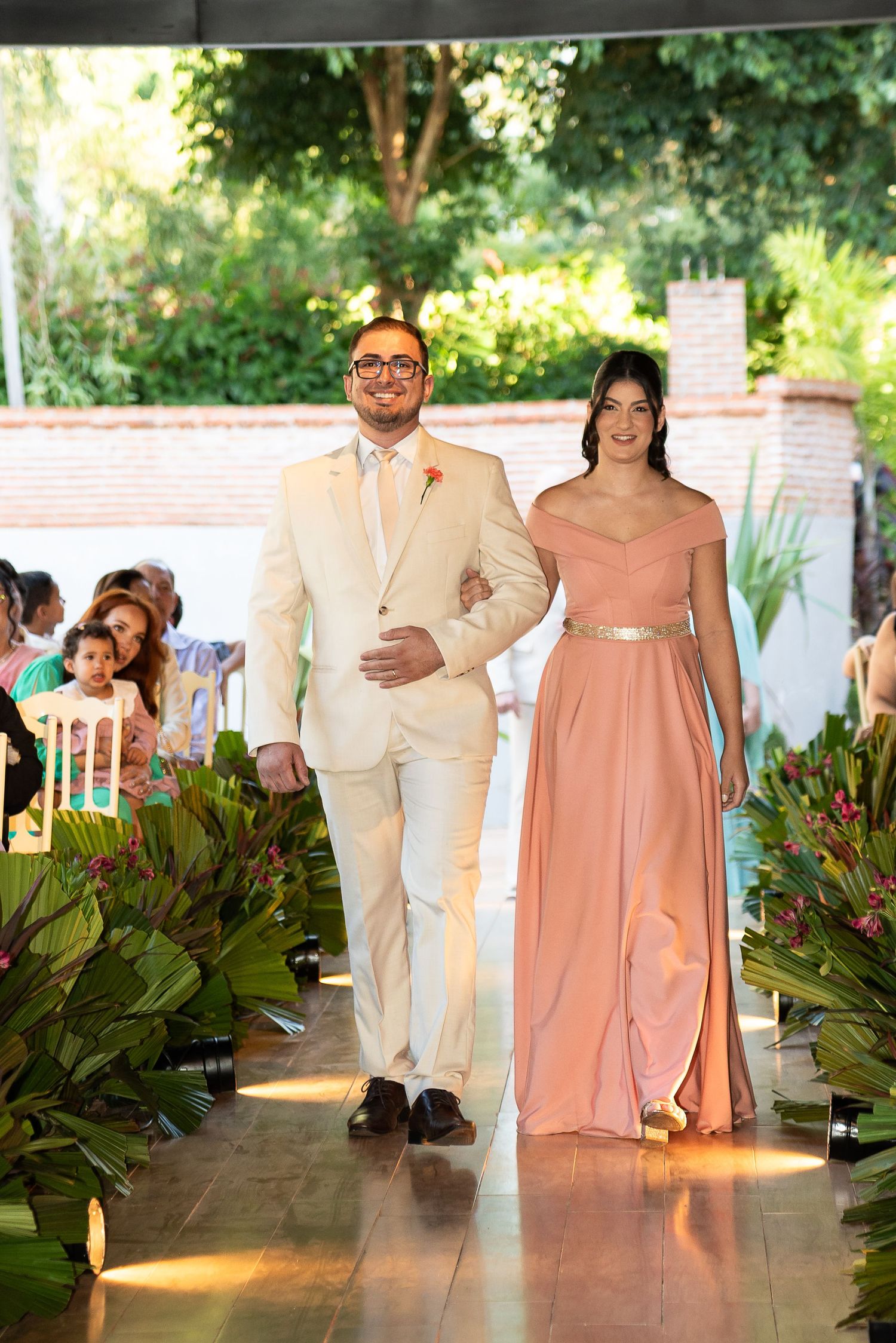 A man in a cream suit and woman in a pink off-shoulder gown walk down a floral-lined wedding aisle.