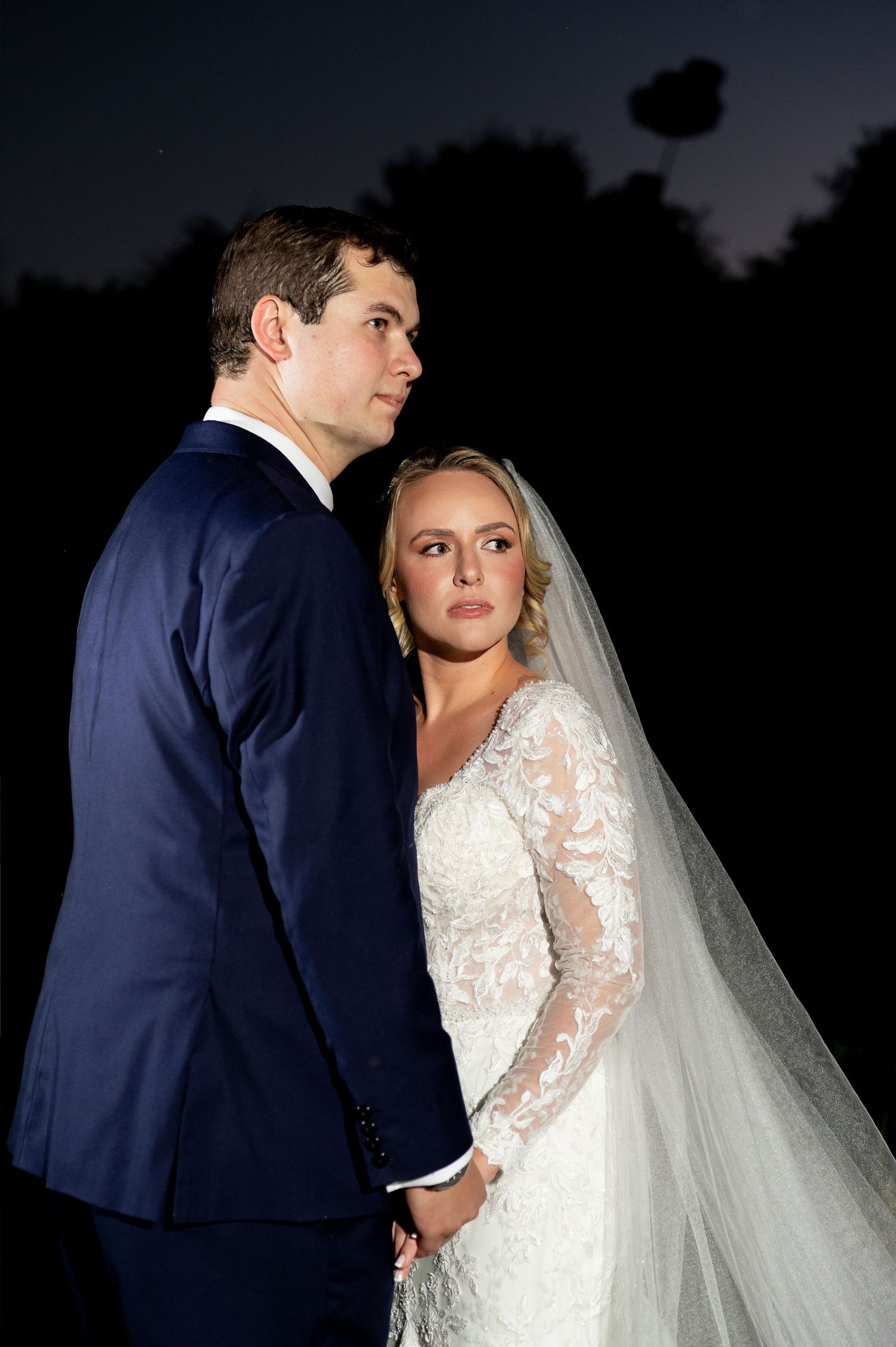 Bride in lace gown and veil with groom in navy suit posing together outdoors at night for wedding portrait.