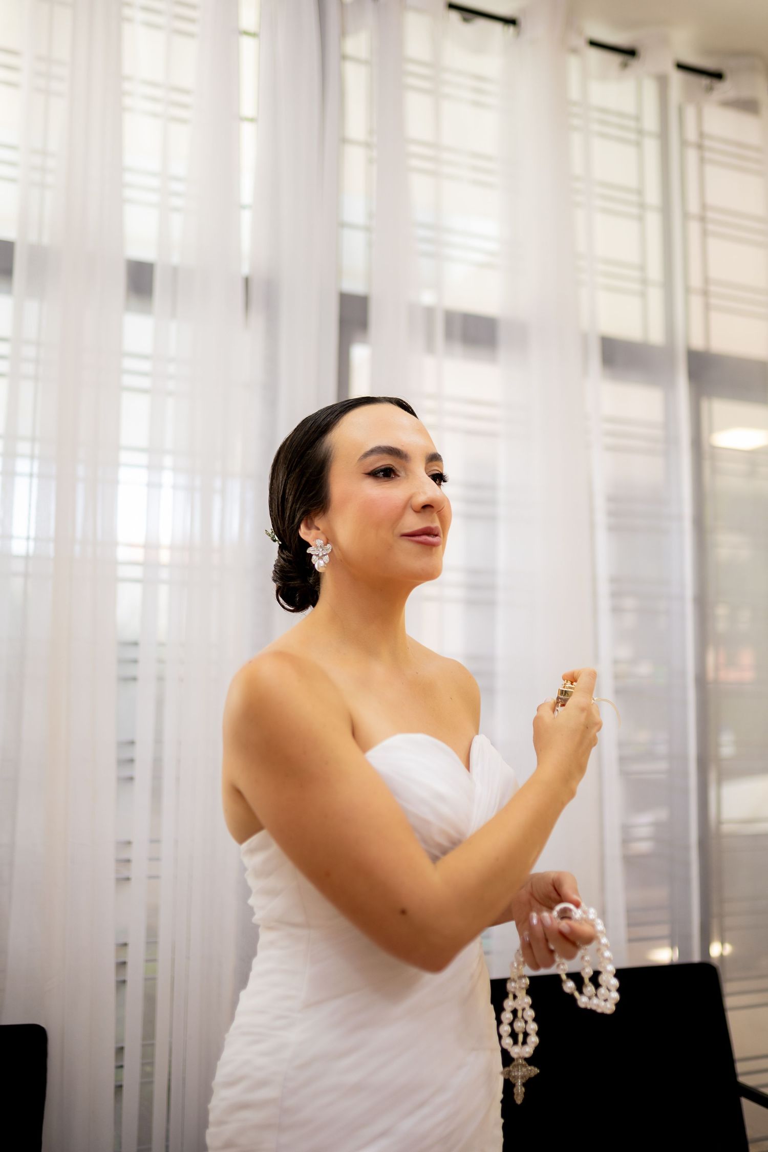 Bride in white strapless gown applying perfume, wearing pearl earrings, with sheer curtains in background.