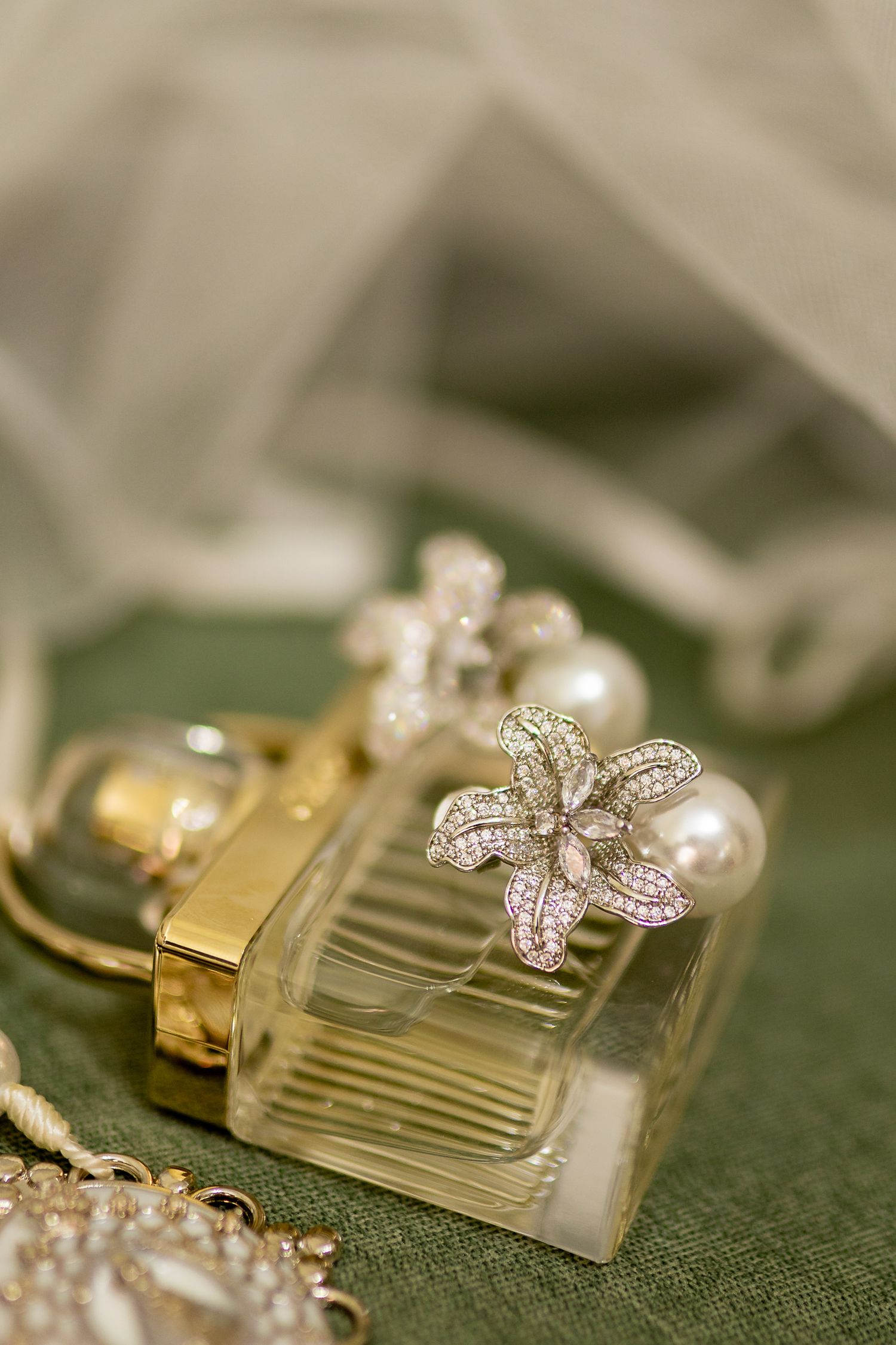 Elegant crystal perfume bottle adorned with pearl and diamond flower earrings on green fabric with sheer ribbon.