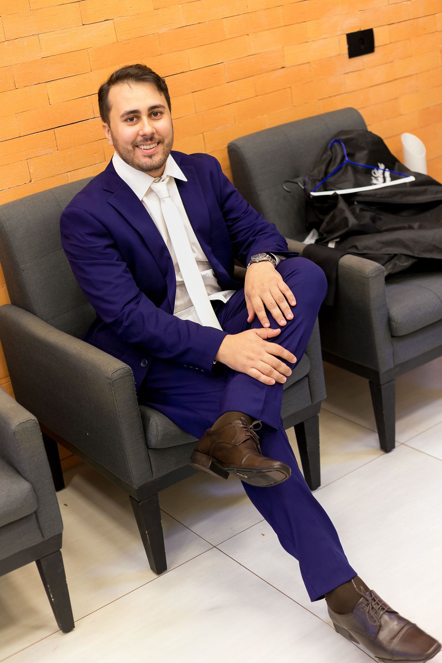 Man in blue suit sitting relaxed in gray chair, smiling, wearing brown dress shoes, brick wall background.