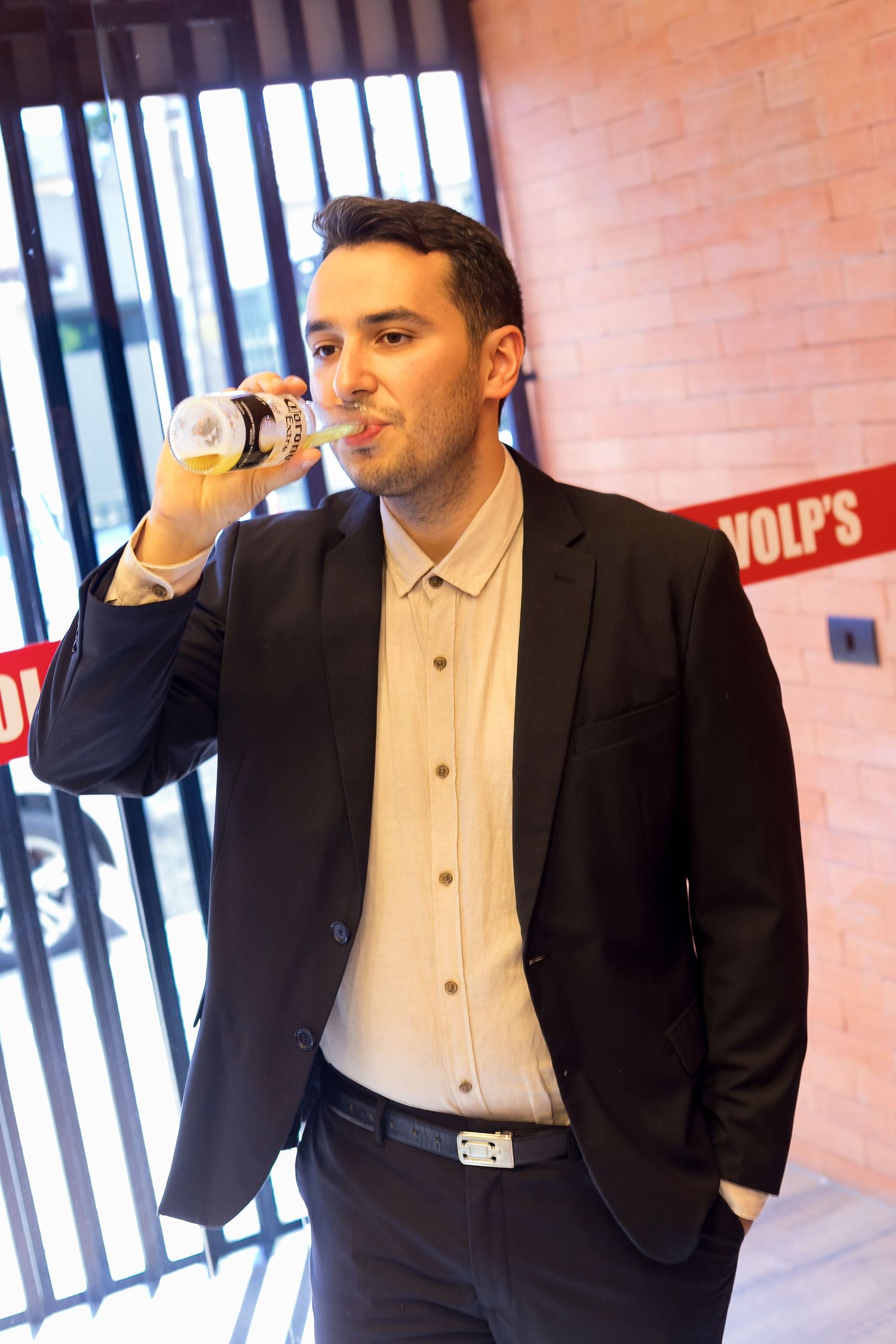 A man in a dark blazer drinks from a can near a brick wall with partial red signage visible in the background.