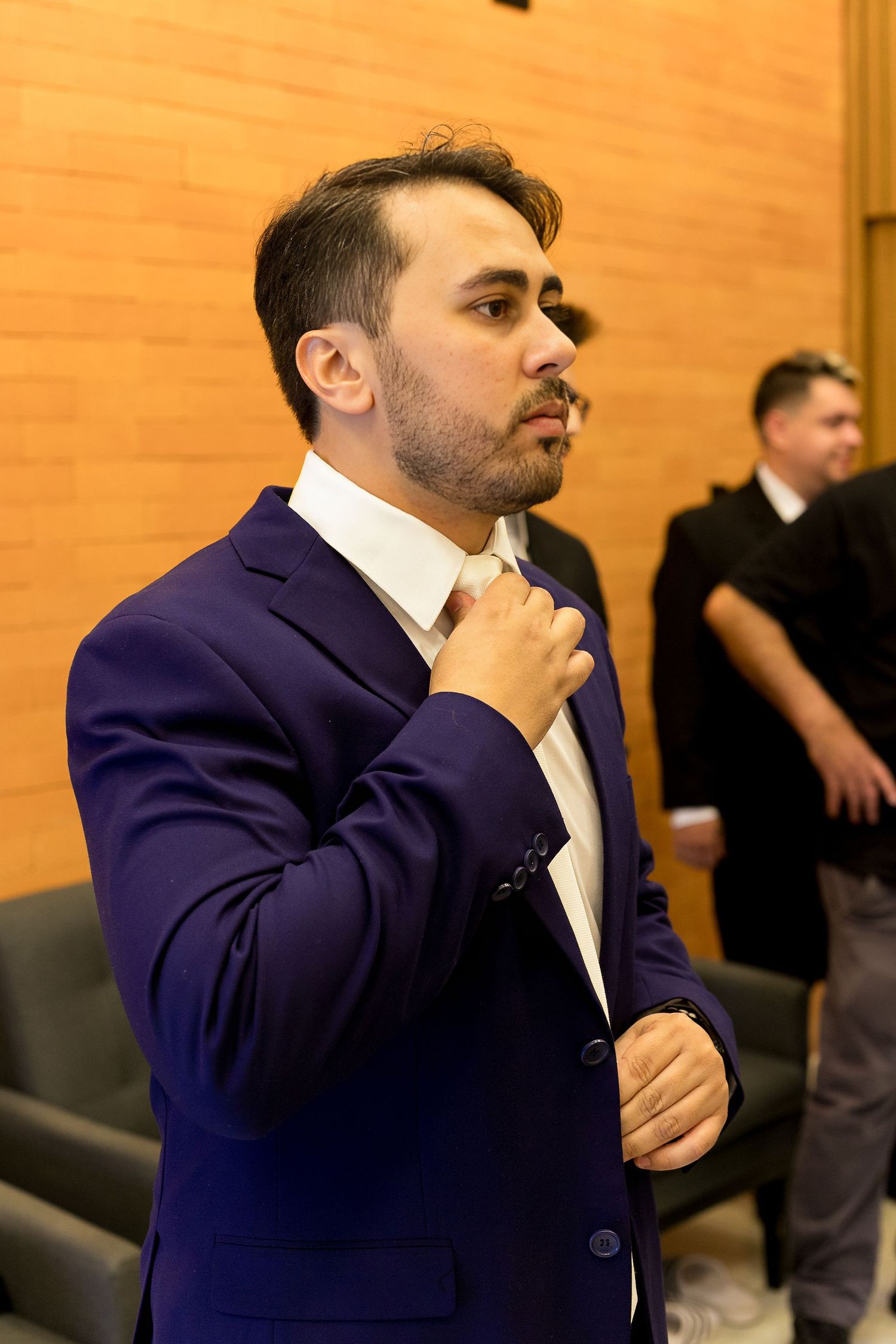 A man in a blue suit adjusts his tie in a formal venue with another person visible in the background.
