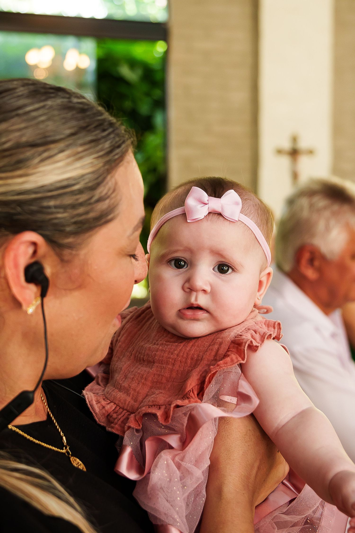 Woman holding a baby girl in pink dress and bow headband at a church baptism ceremony.