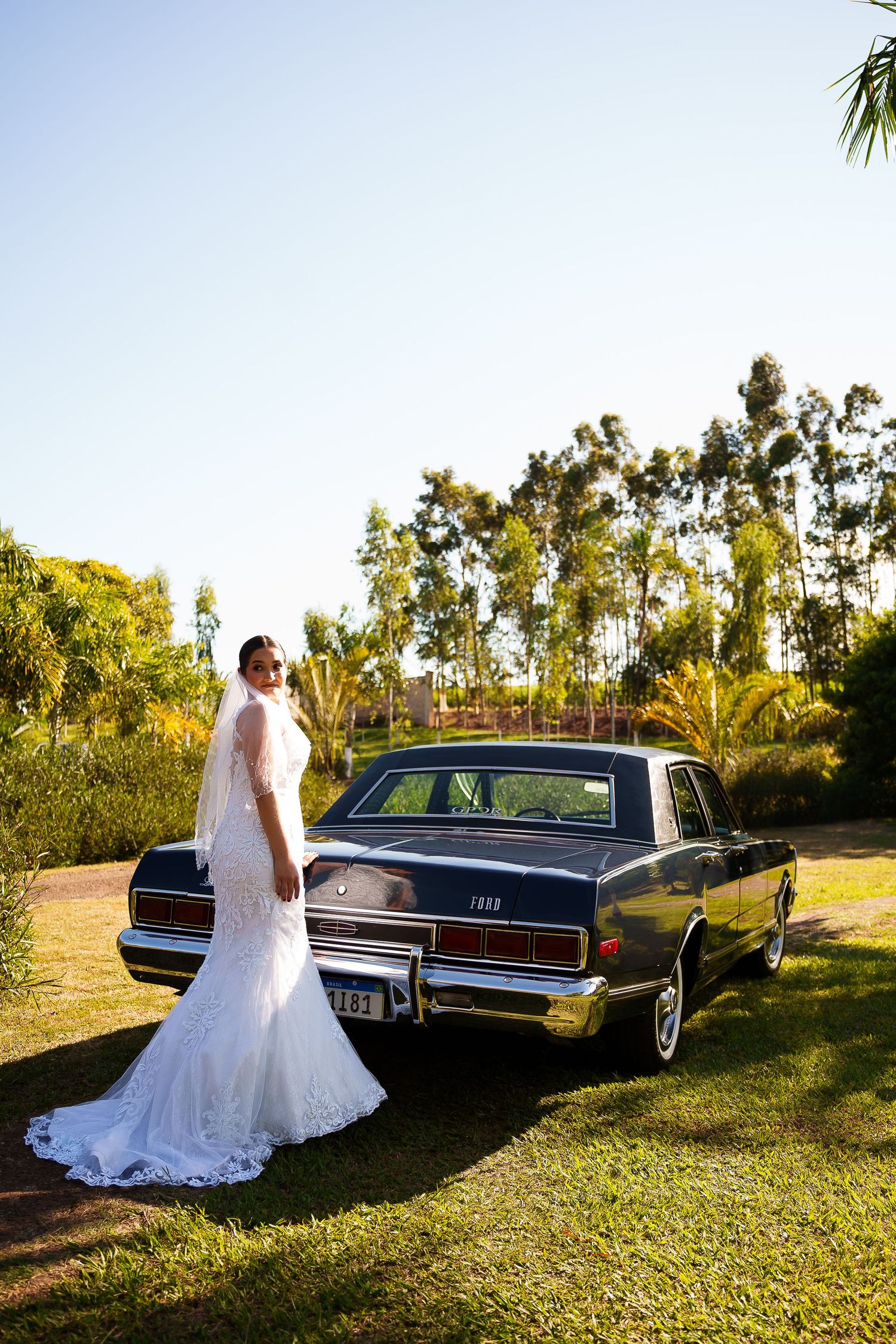 Bride in white lace gown and veil poses beside a classic black car on green lawn with tropical trees backdrop.