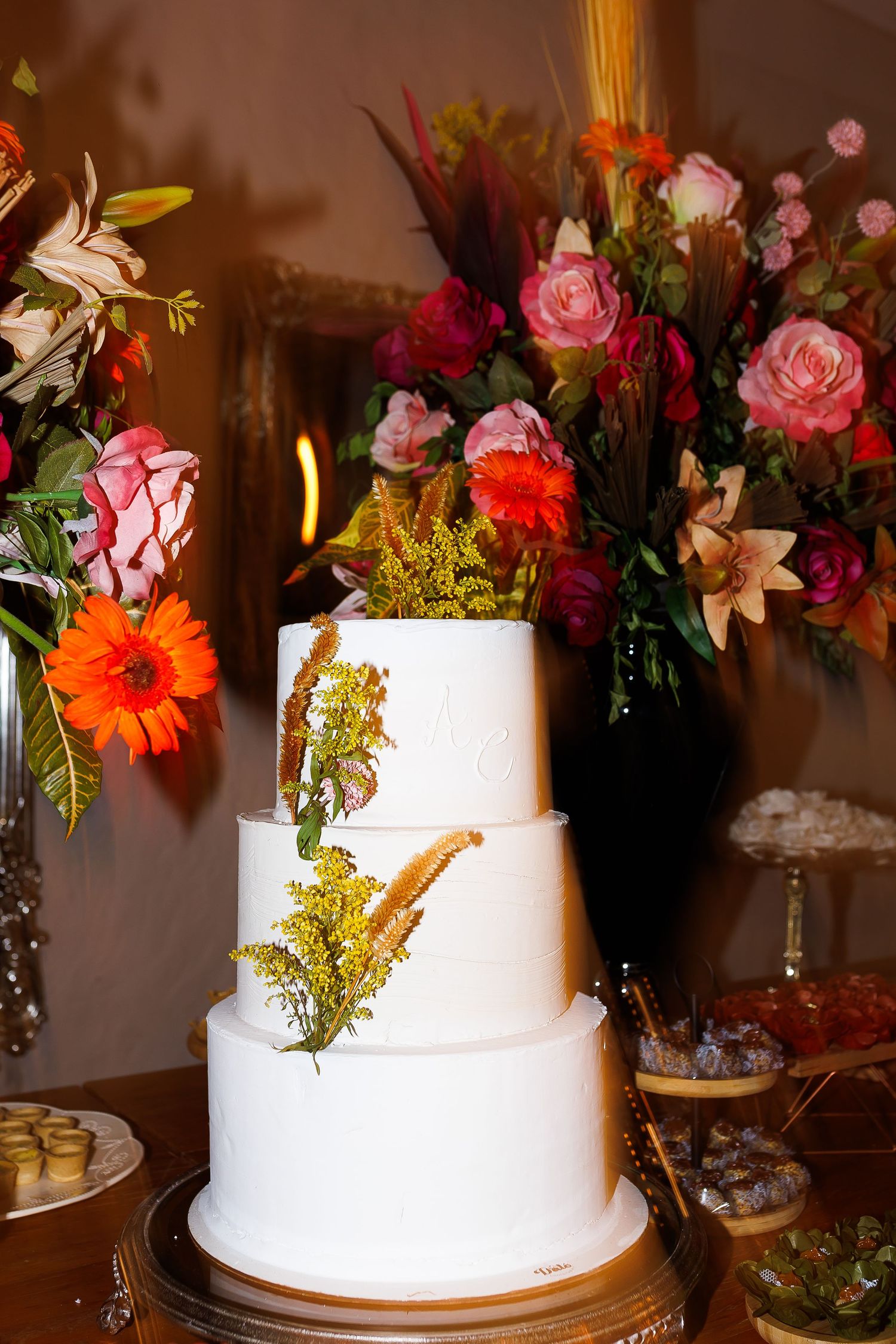 Three-tier white wedding cake with golden botanical accents, surrounded by vibrant floral arrangements in pink, orange, and red.