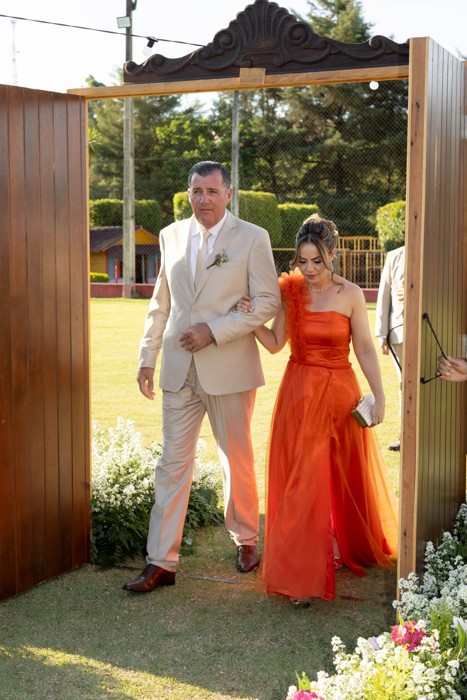 Man in beige suit escorts woman in orange gown through wooden doors at outdoor wedding ceremony.