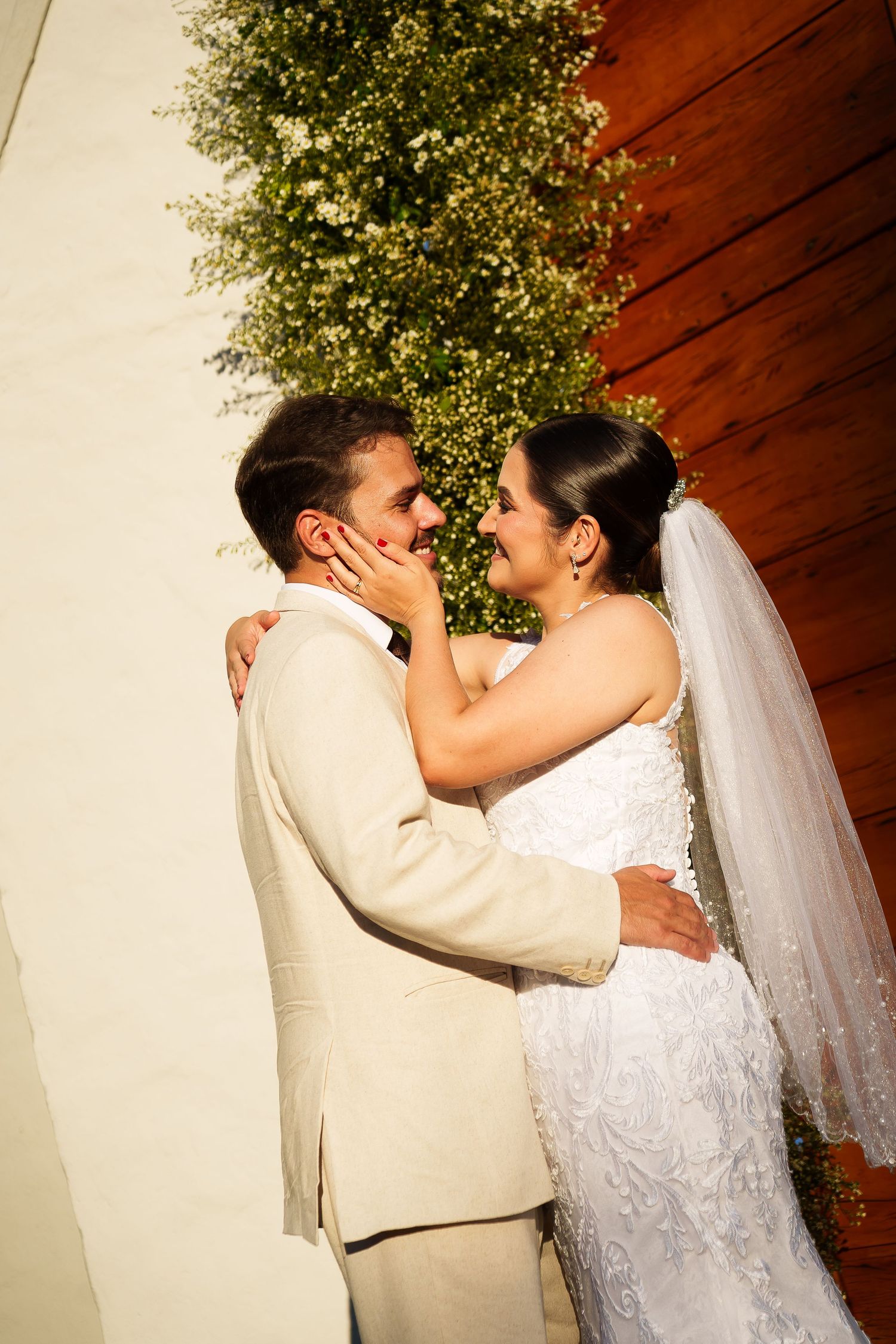 Bride in white gown and veil embraces groom in beige suit, sharing a romantic moment outdoors near greenery.