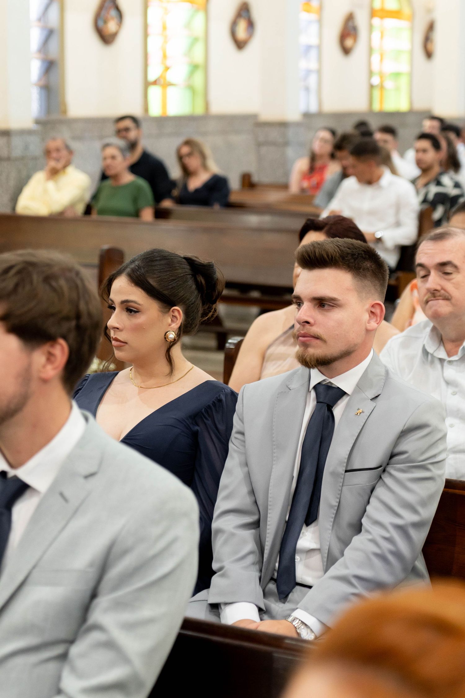 Guests seated in church pews during a wedding ceremony, woman in navy dress beside man in gray suit.