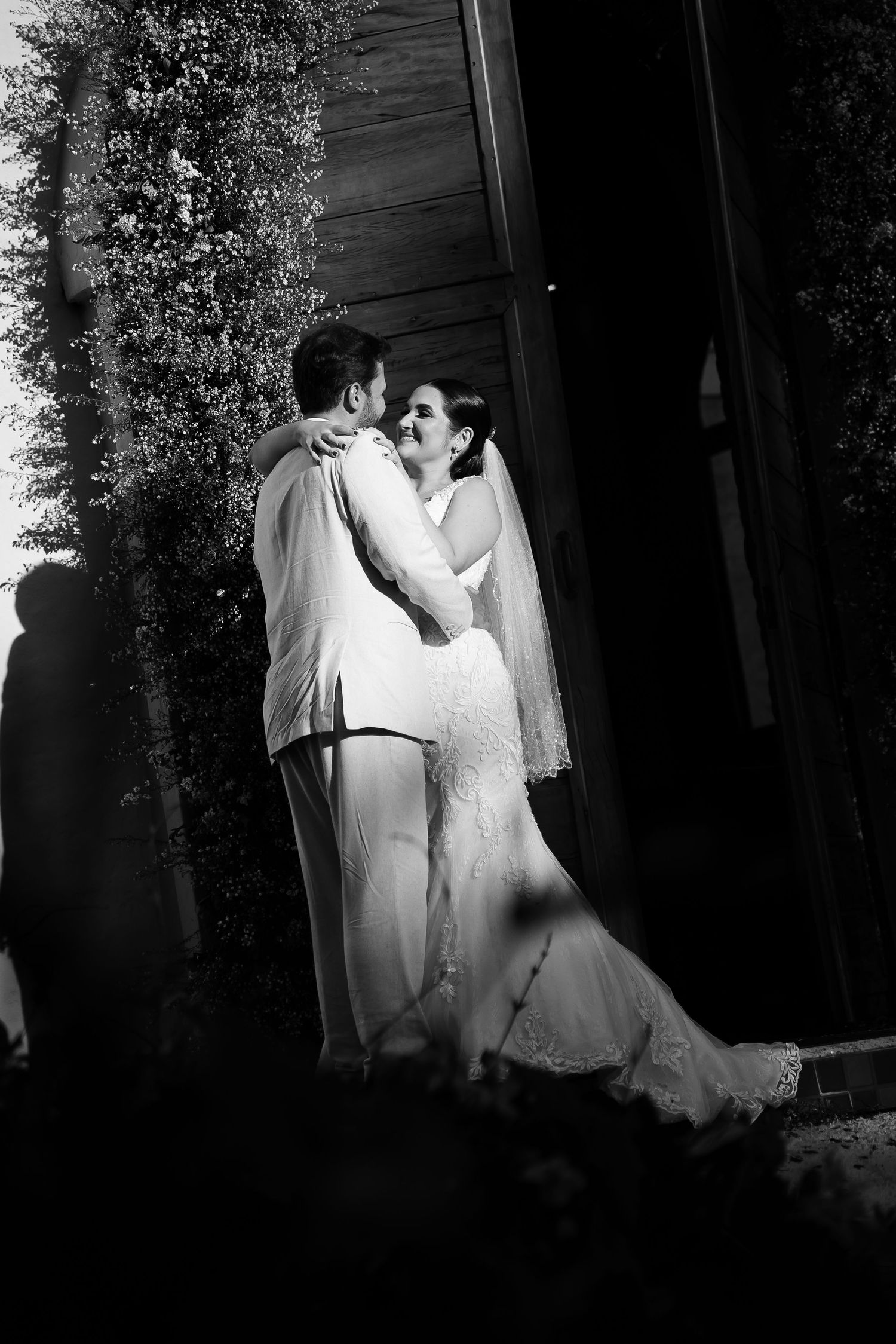 Black and white wedding photo of bride and groom embracing outdoors near lush greenery.