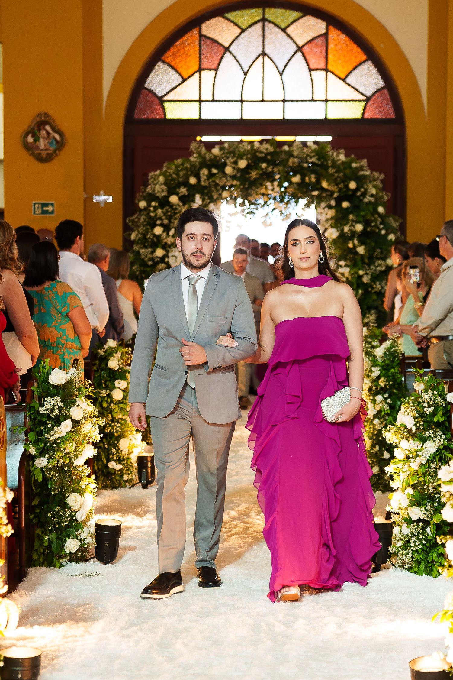 Couple walks down floral wedding aisle, man in gray suit, woman in magenta gown, church ceremony.