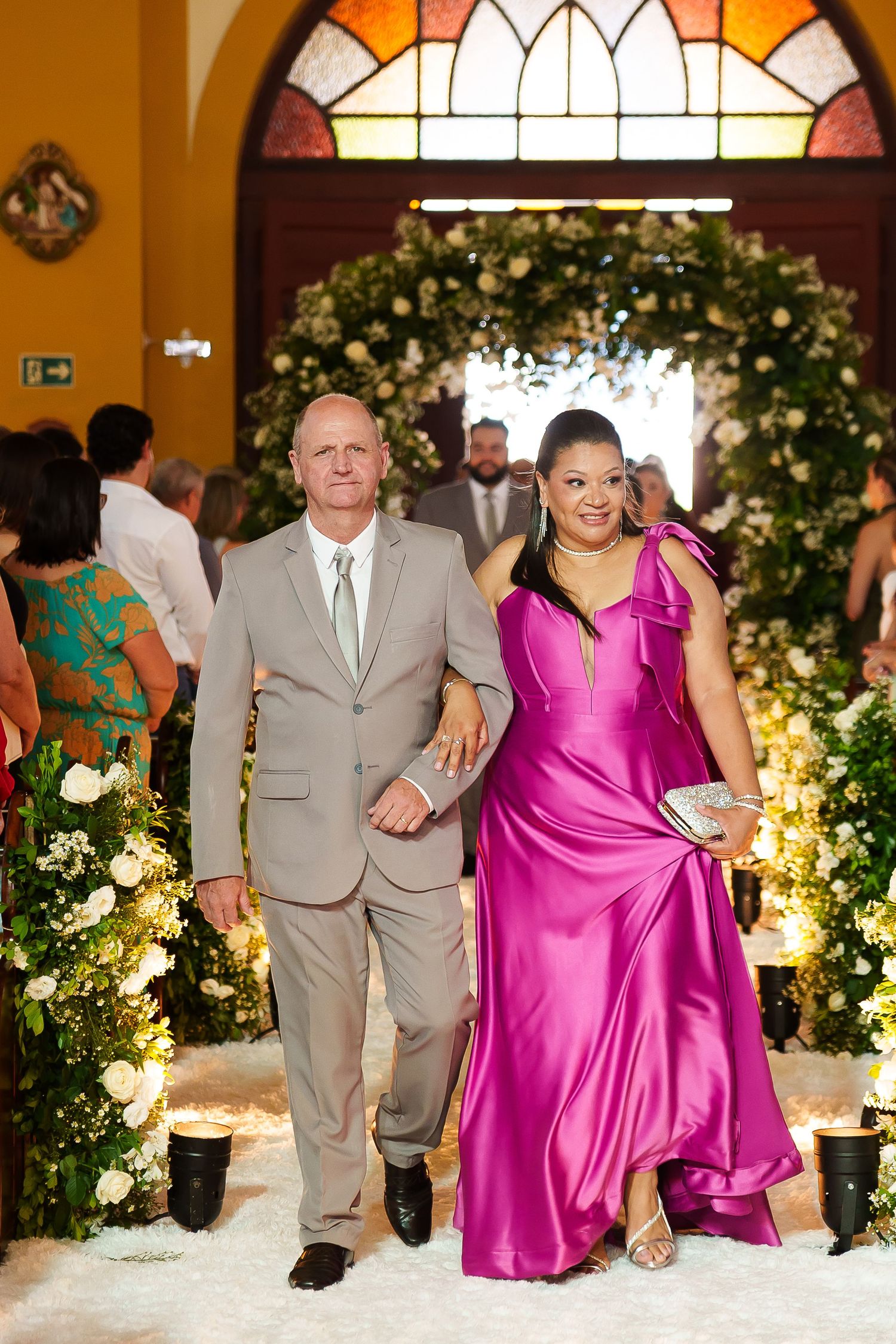 Couple walks down floral wedding aisle, man in tan suit, woman in magenta gown, church setting.