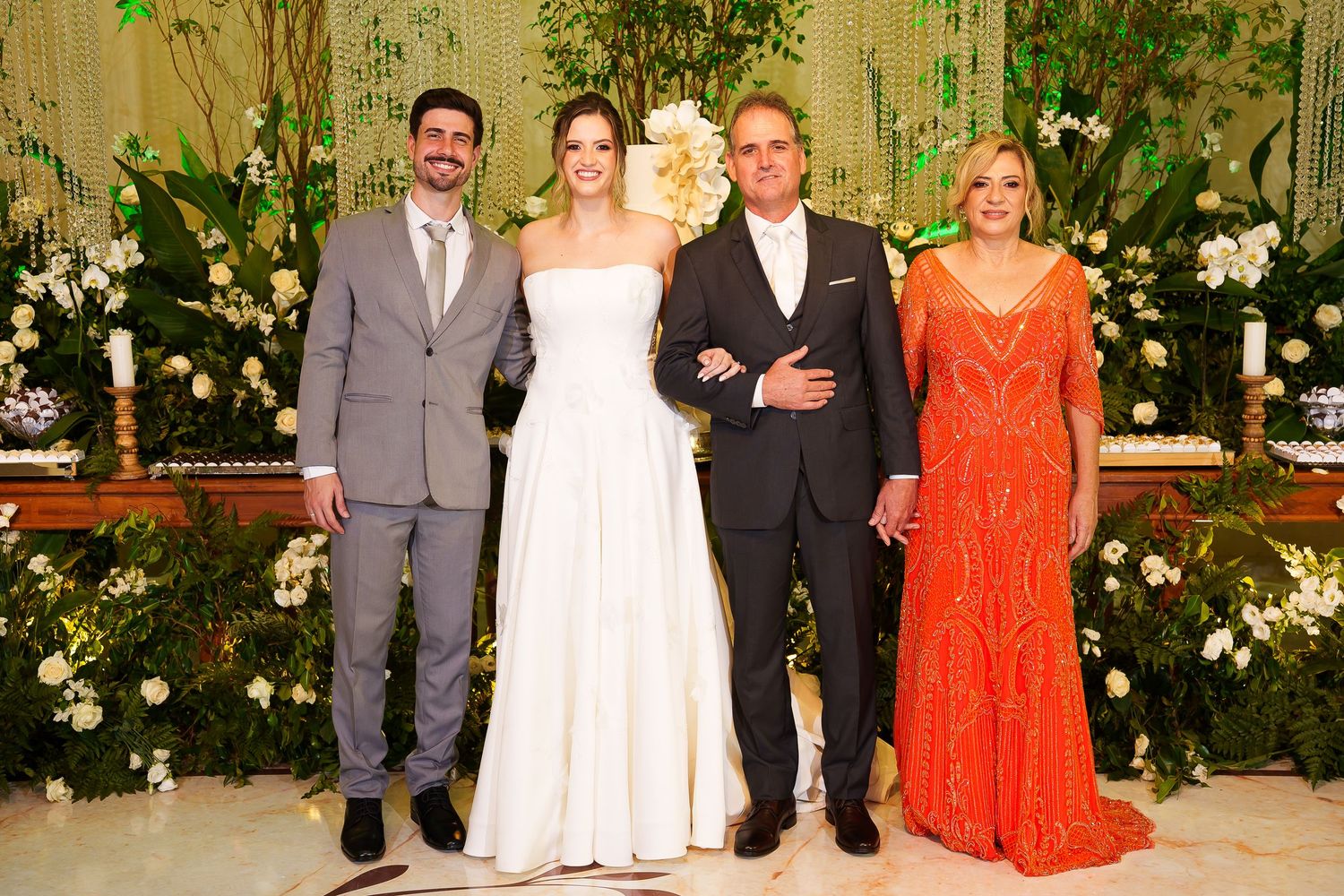Elegant wedding party with bride in white gown, groom in suit, and guests in formal attire posing before floral backdrop.