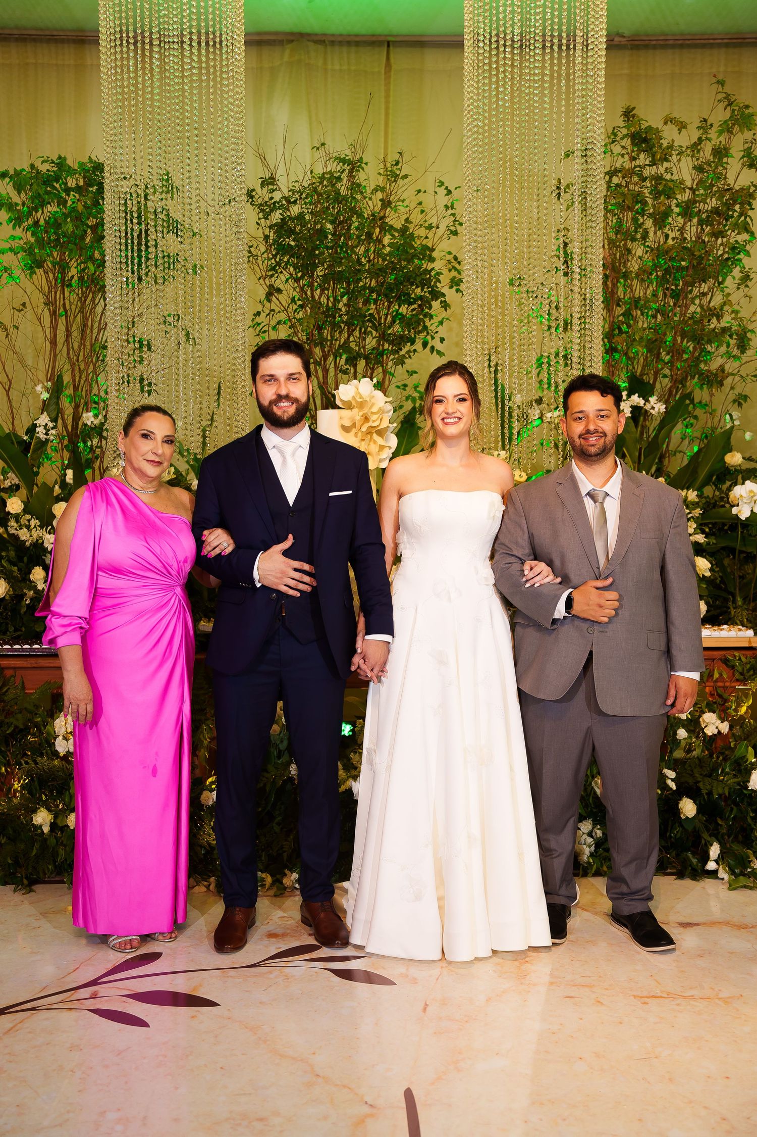 Wedding party poses with bride in white gown and groom in navy suit, flanked by guests in pink dress and gray suit.