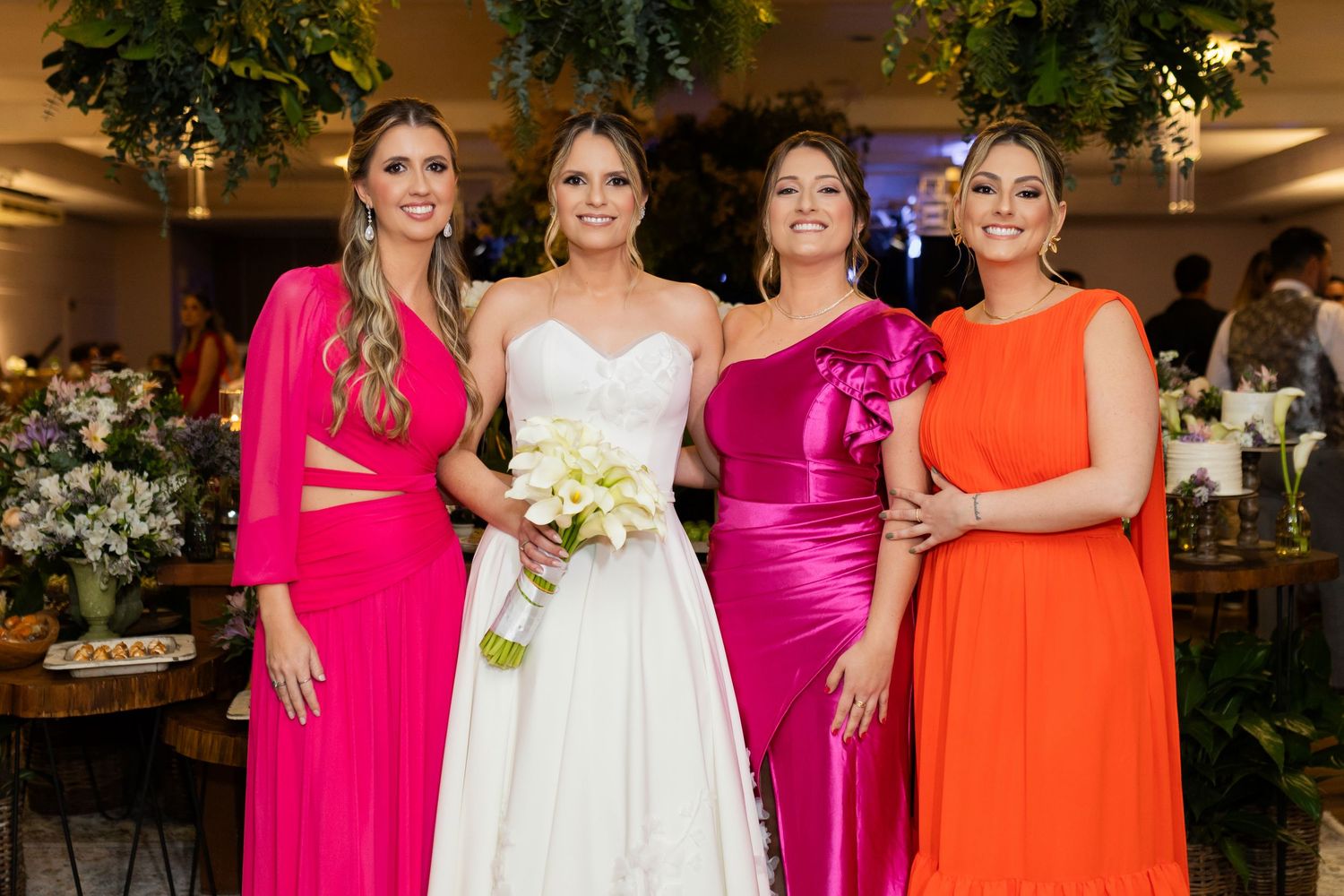 Bride in white gown poses with three bridesmaids in vibrant pink and orange dresses at elegant wedding reception.