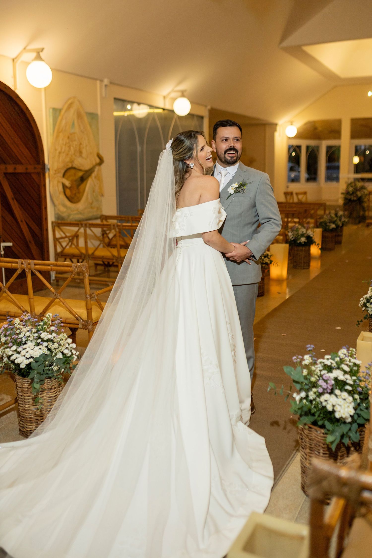 Bride in white off-shoulder gown with veil and groom in gray suit pose together in a warmly lit chapel.