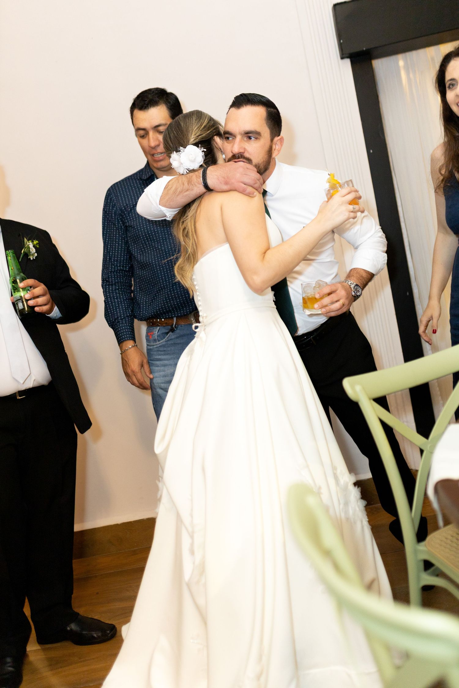 Bride and groom share a warm embrace during their wedding reception, surrounded by guests in a bright venue.