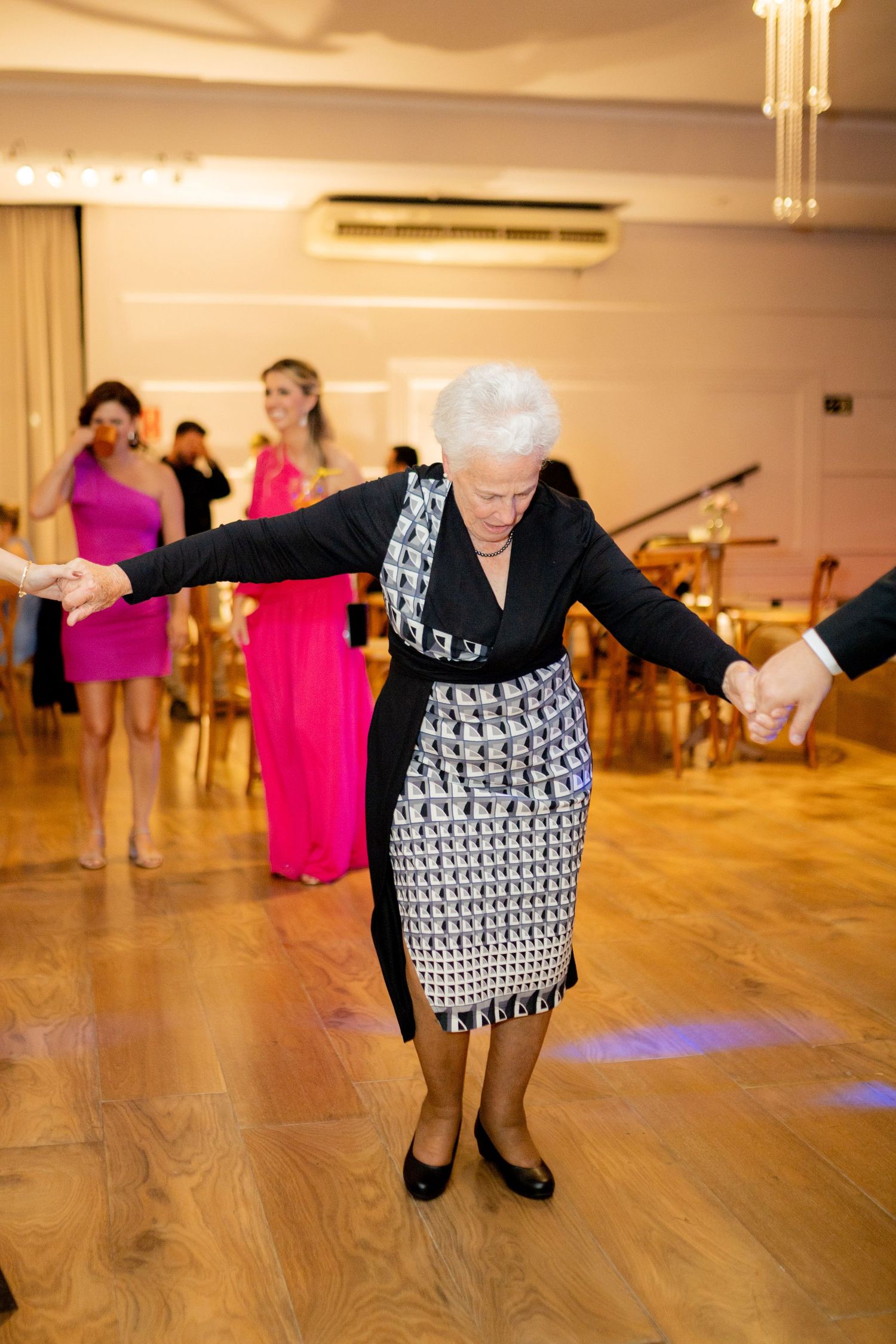 Elderly woman dancing at a formal event, arms outstretched, wearing a patterned dress and black cardigan on a wooden dance floor.