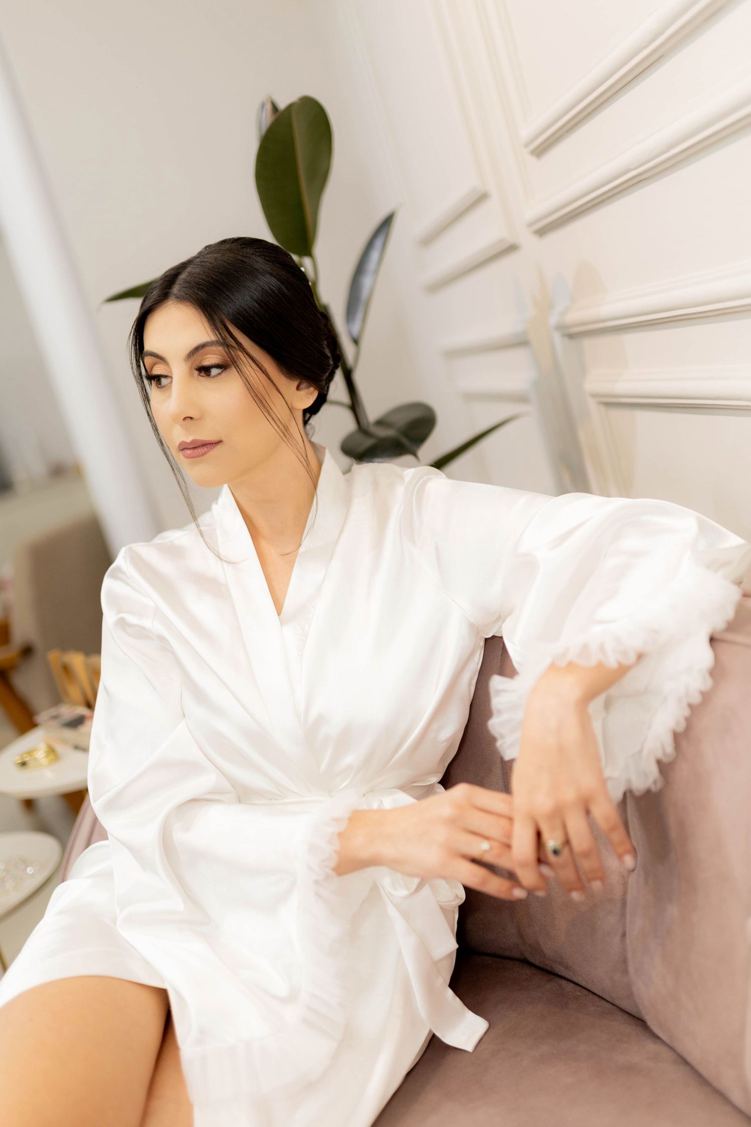 A woman in a white satin robe with feather trim sits gracefully on a pink sofa in an elegant room.