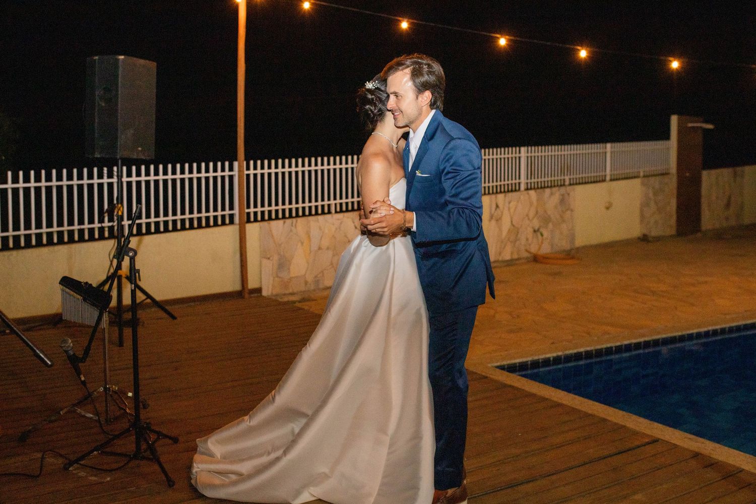 Bride and groom share their first dance outdoors at night beside a pool, surrounded by warm string lights.