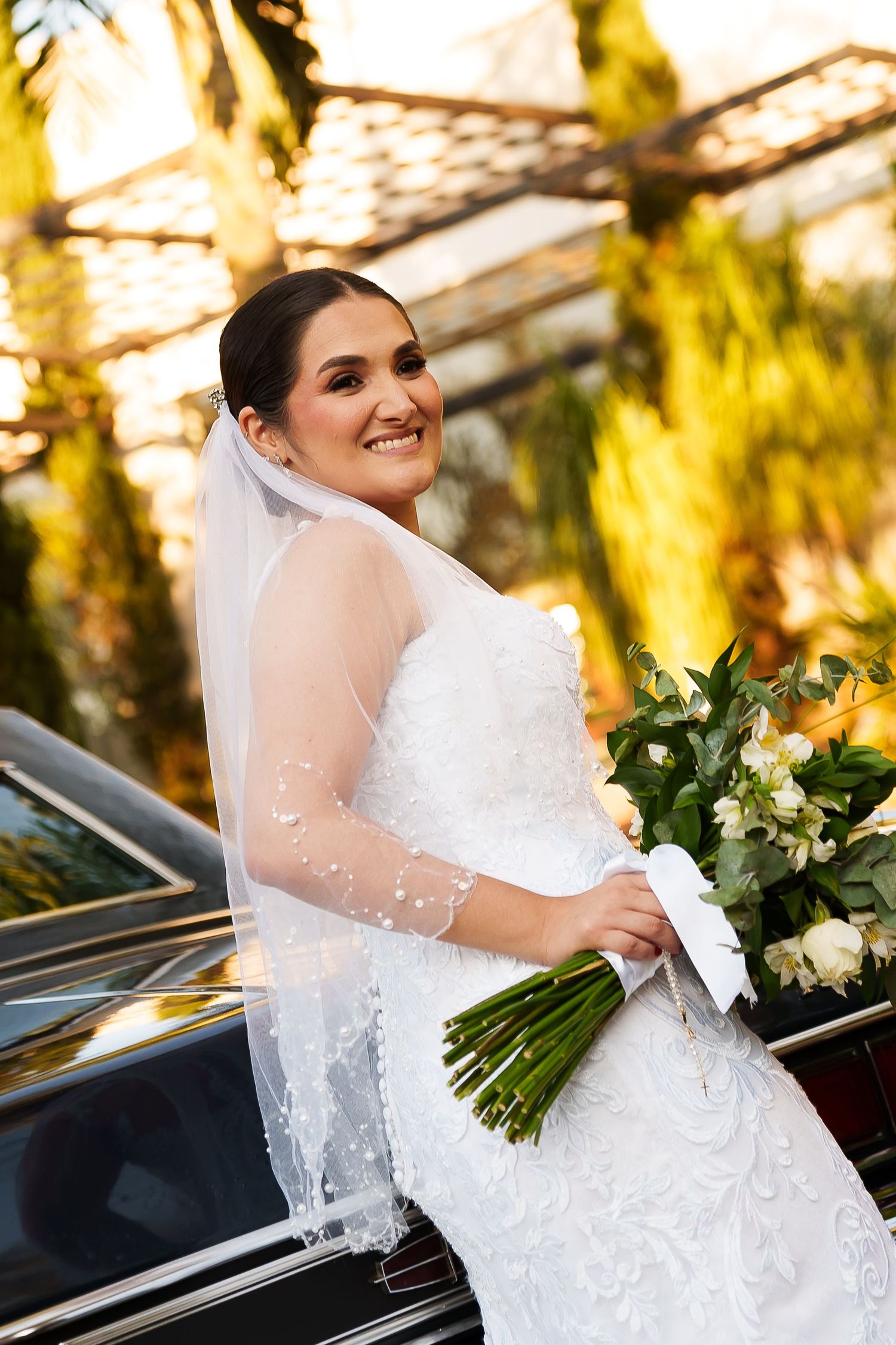 Smiling bride in white lace gown and veil holds bouquet of white flowers beside a car with green trees behind.