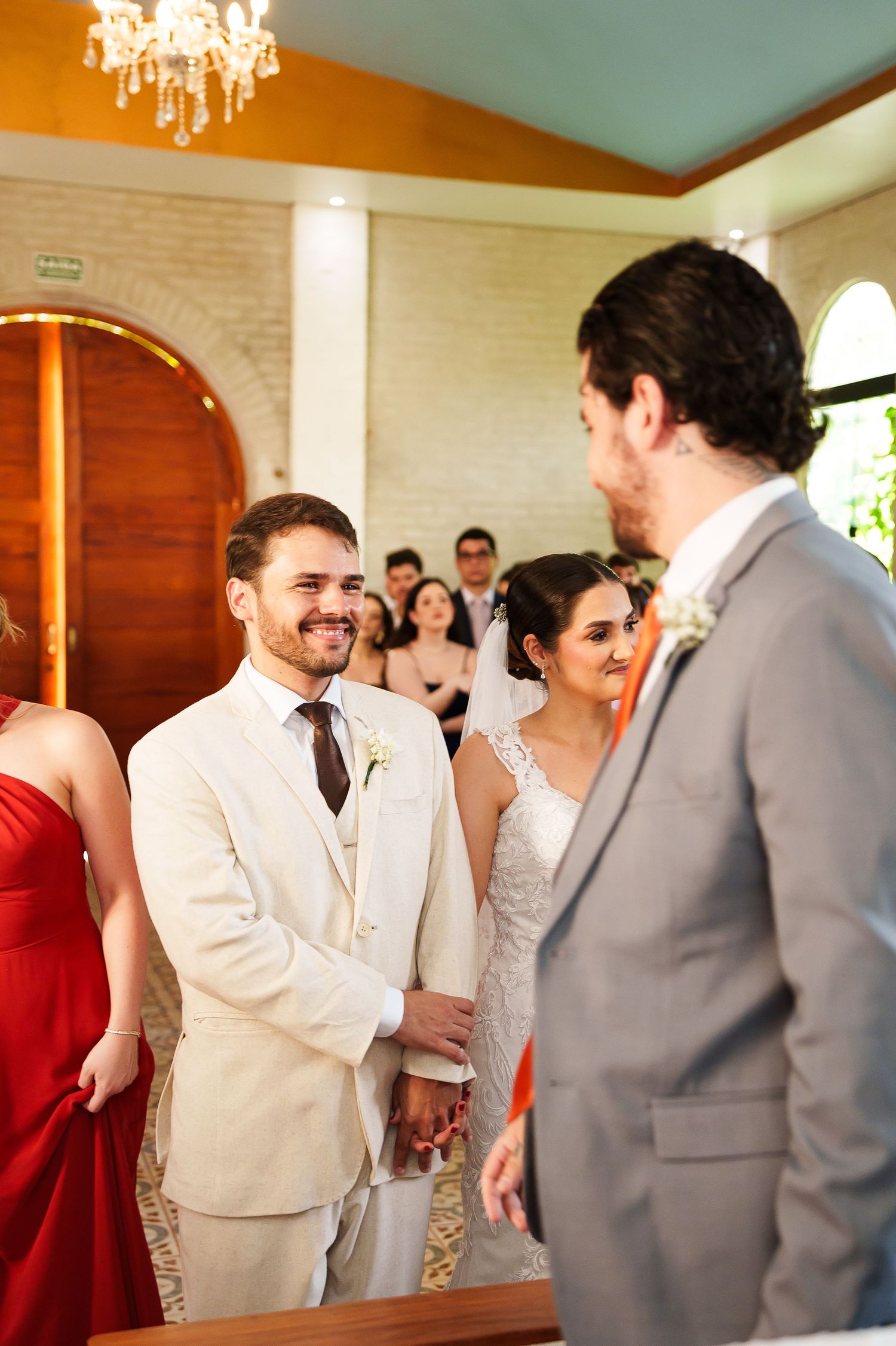 Groom in cream suit smiling during wedding ceremony at elegant church with bridal party in background.