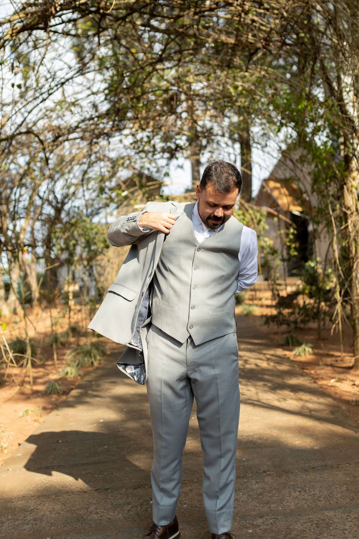 Man in gray suit vest putting on jacket outdoors in garden path with trees overhead.