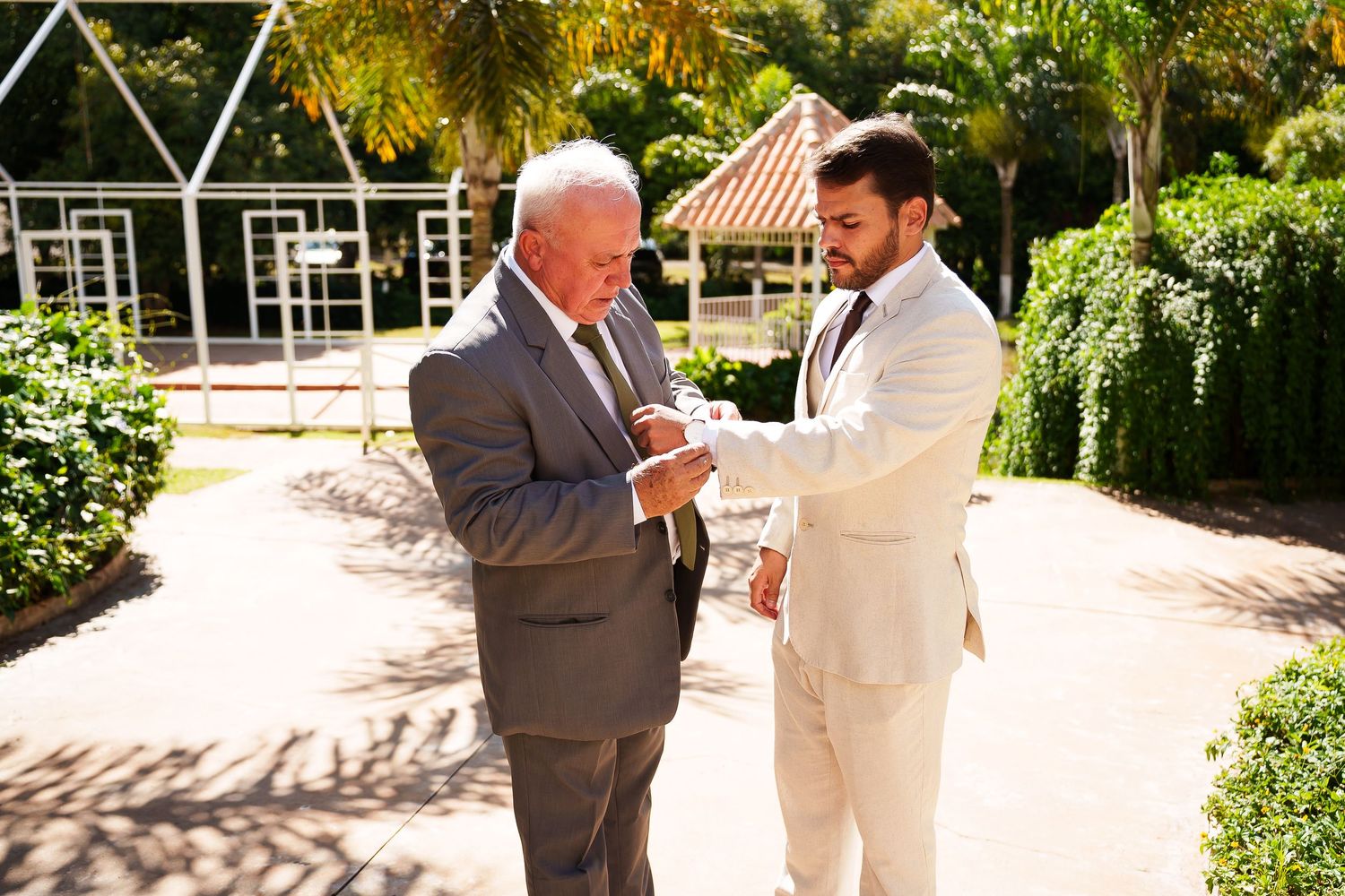 Groom in beige suit adjusting older mans gray suit jacket outdoors at wedding venue with tropical greenery.