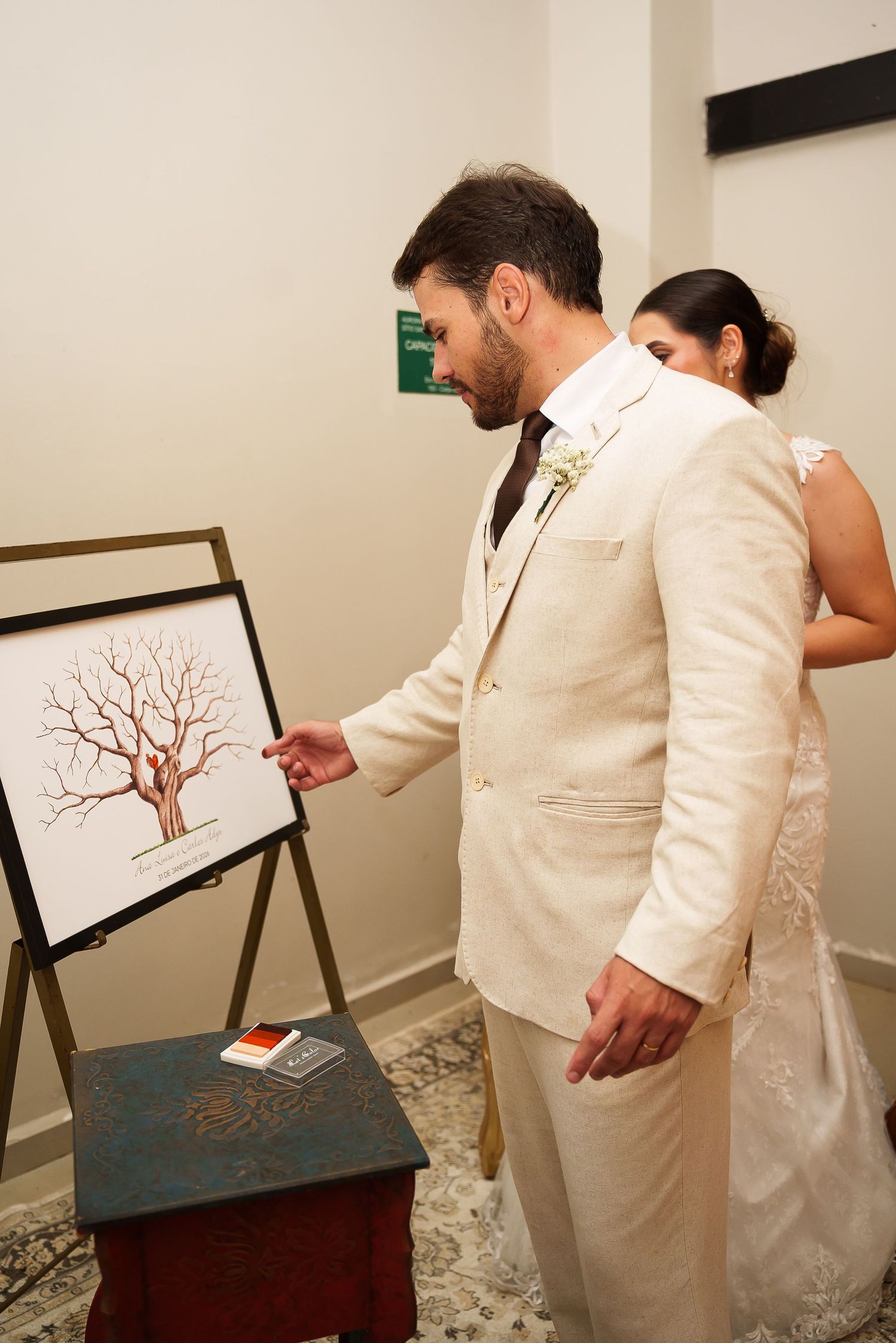 Groom in cream suit points at fingerprint tree wedding guest book on easel, bride in white lace dress stands behind.