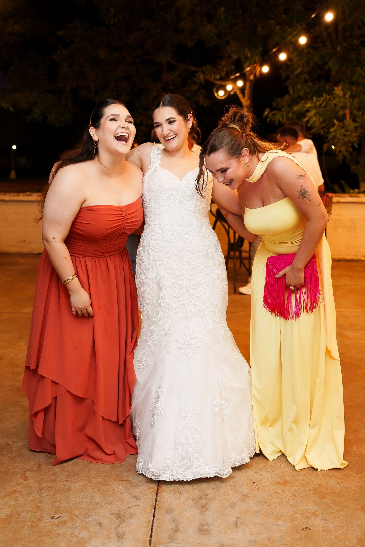 Bride in white lace gown laughing with bridesmaids in red and yellow dresses at outdoor wedding reception.