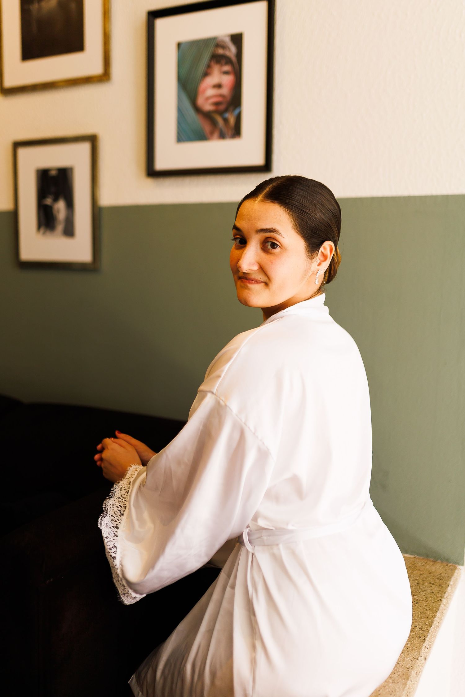 Woman in white lace robe sitting and looking over shoulder, framed portraits on green wall behind her.