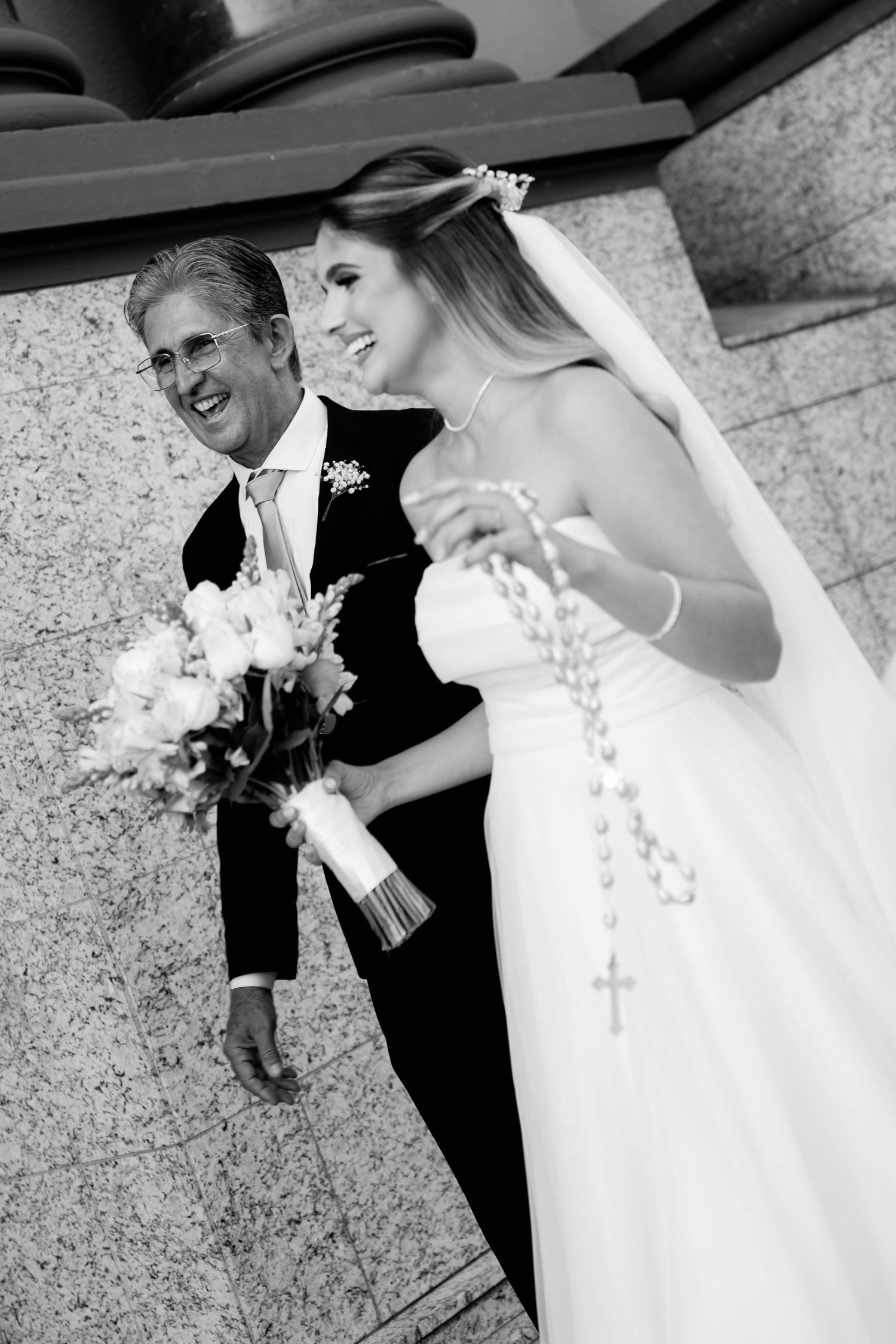 Black and white photo of smiling bride holding rosary and bouquet with groom in tuxedo outside church.