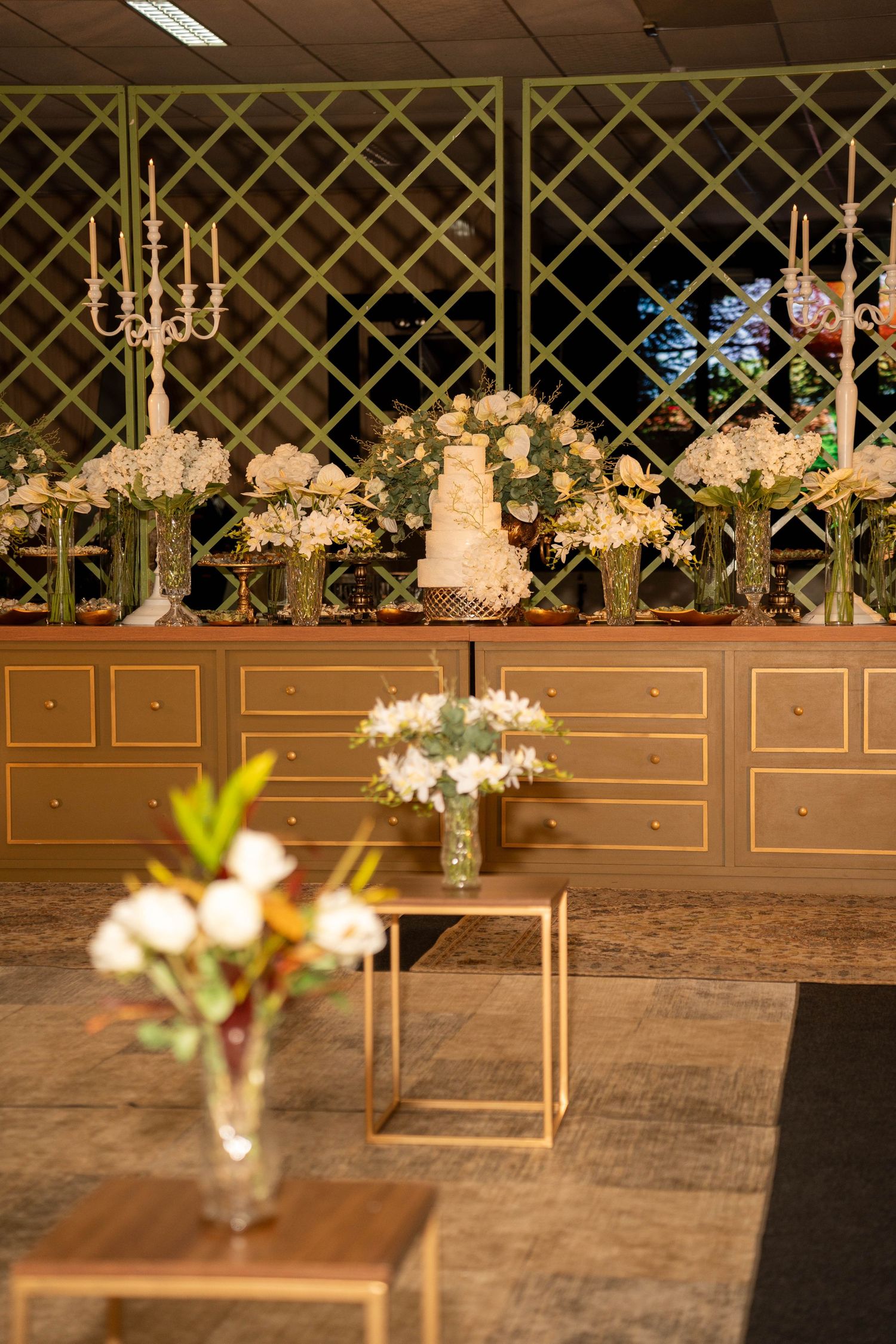 Elegant wedding cake display with white floral arrangements, candelabras, and gold furniture in a decorated reception venue.