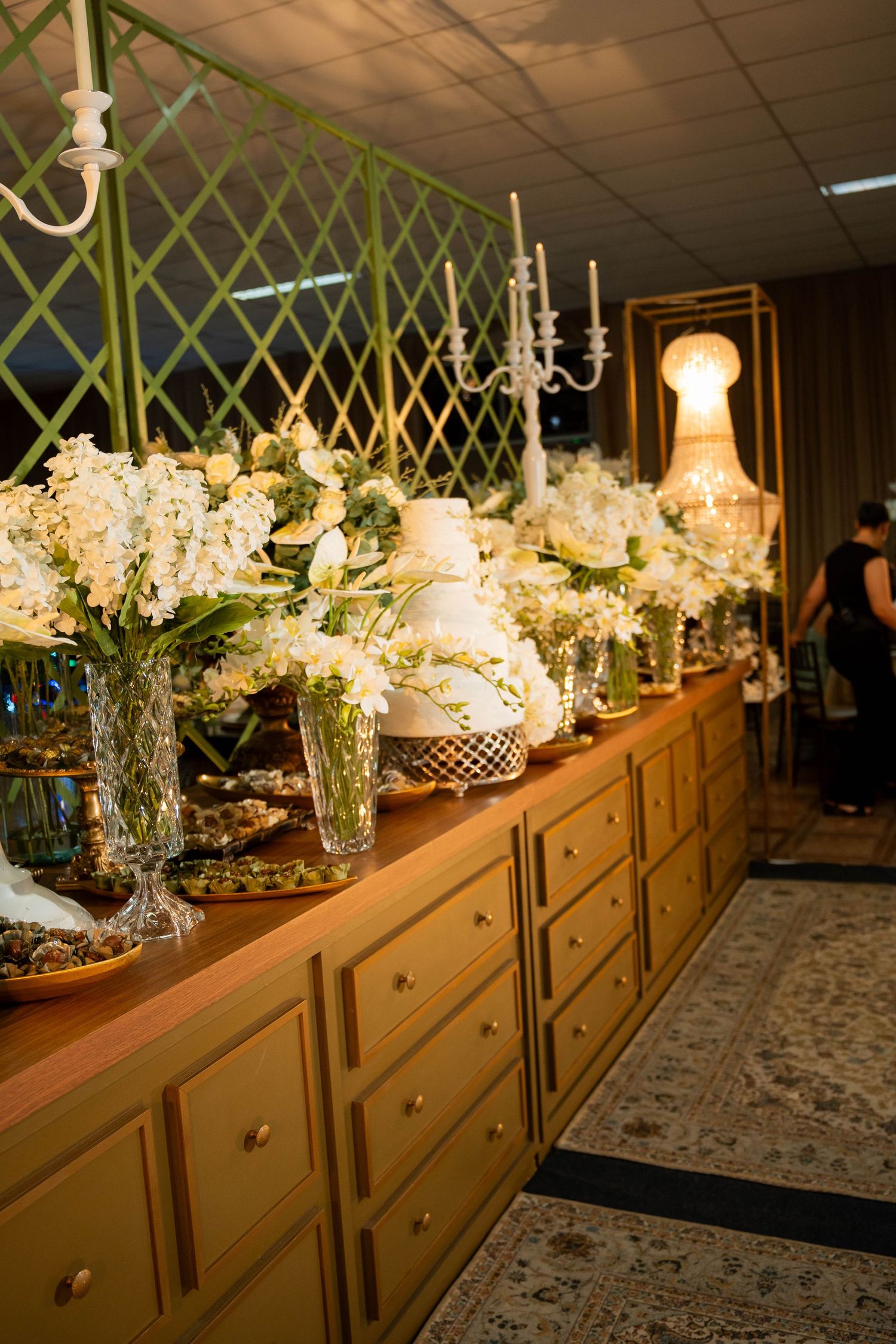 Elegant wedding reception buffet table with white floral arrangements, candelabras, and tiered cake on gold drawers.