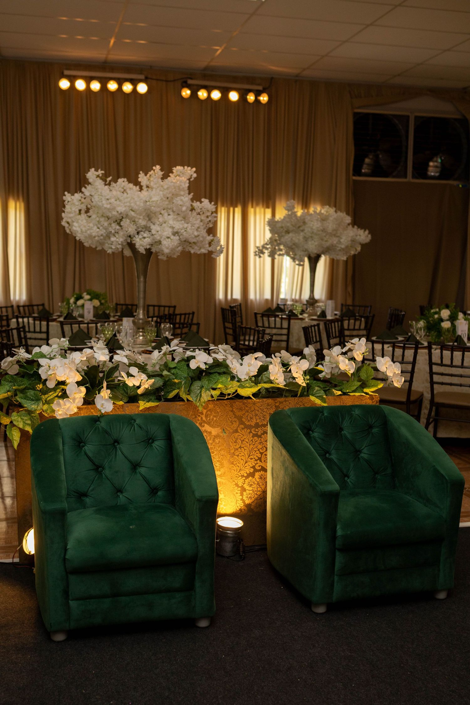 Elegant wedding reception featuring two green velvet tufted chairs beside a gold sweetheart table with white floral arrangements.