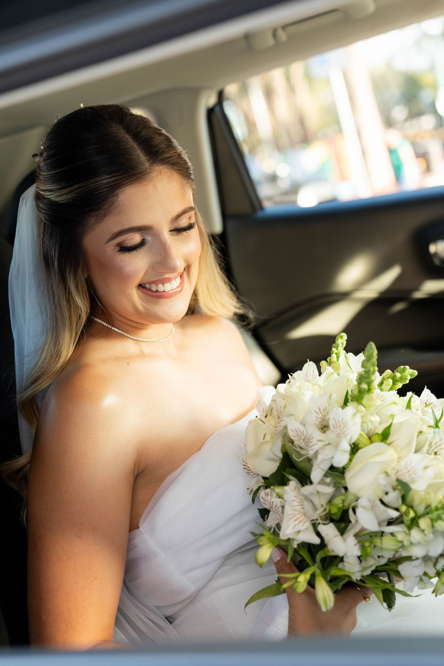 Smiling bride in white dress holding bouquet of white flowers while sitting in a car on her wedding day.