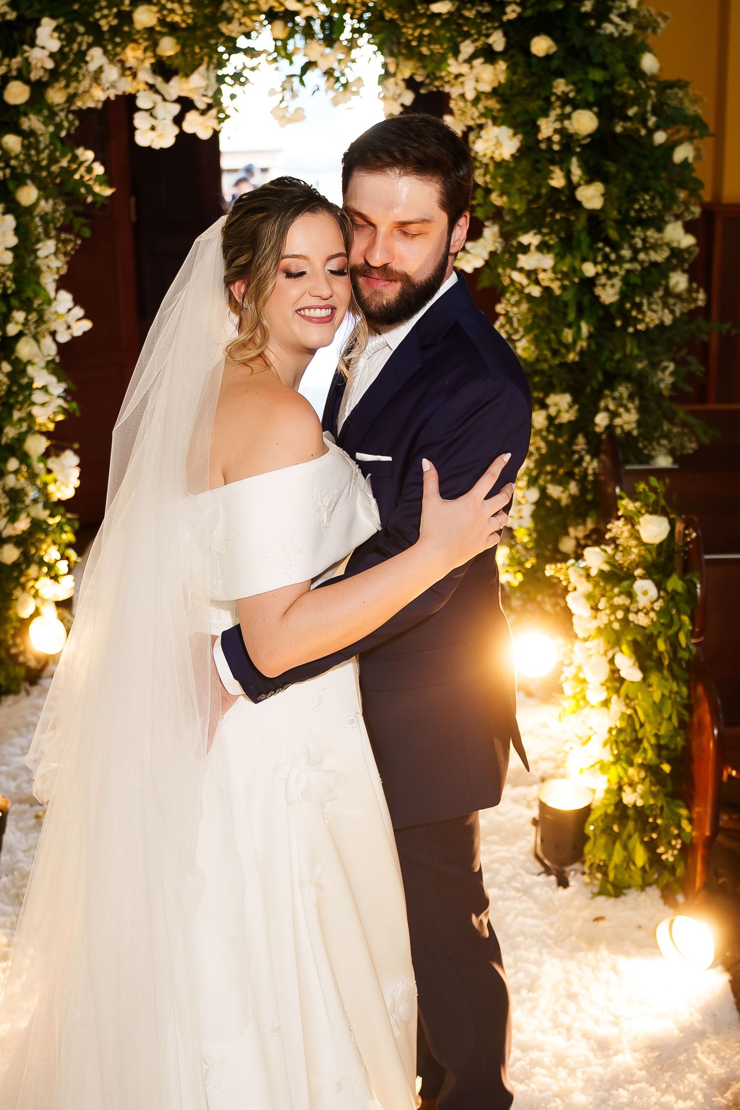 Bride and groom embracing under floral arch with white roses and warm lights at elegant wedding ceremony.