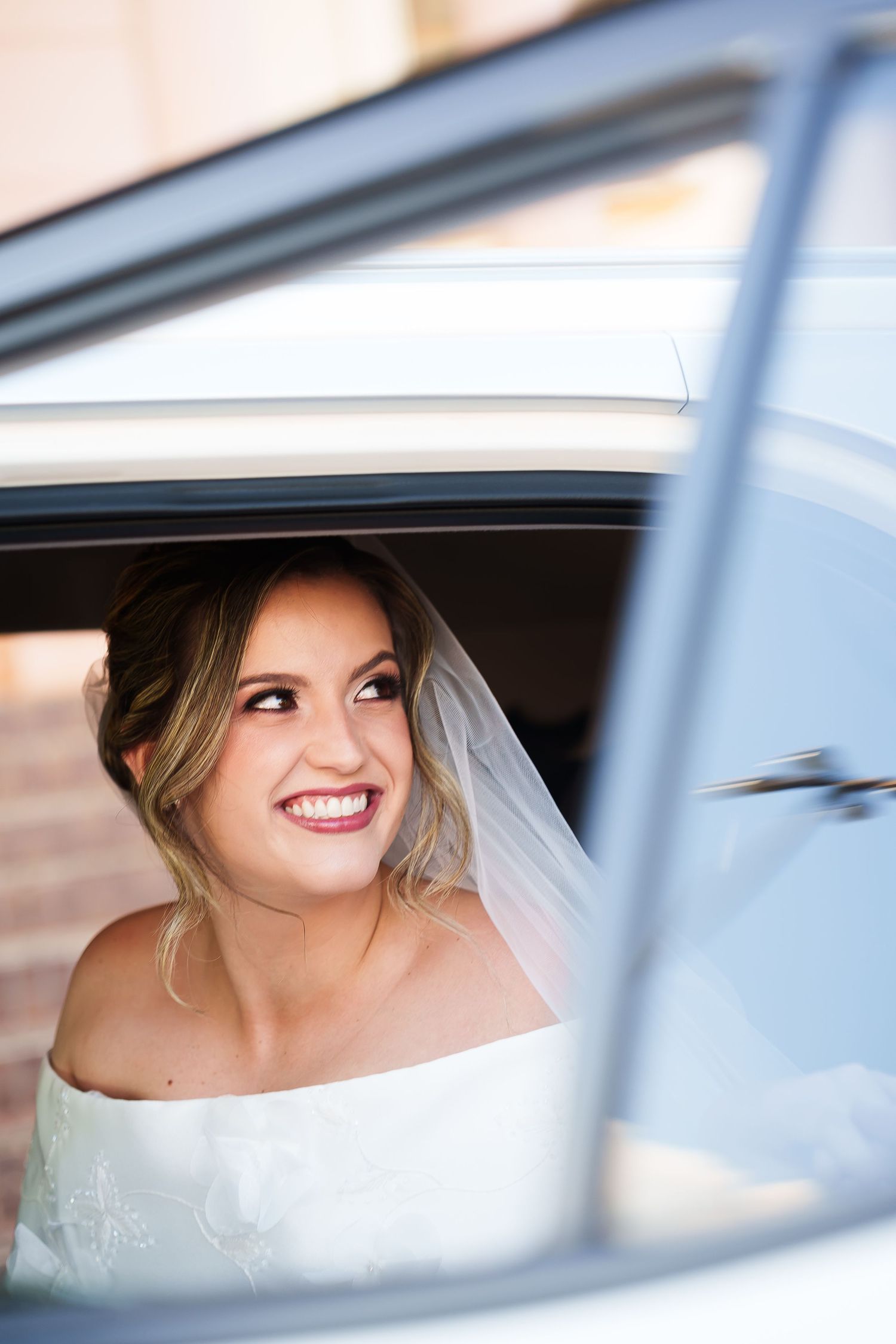 Smiling bride in white dress and veil looking up through open car window on her wedding day.