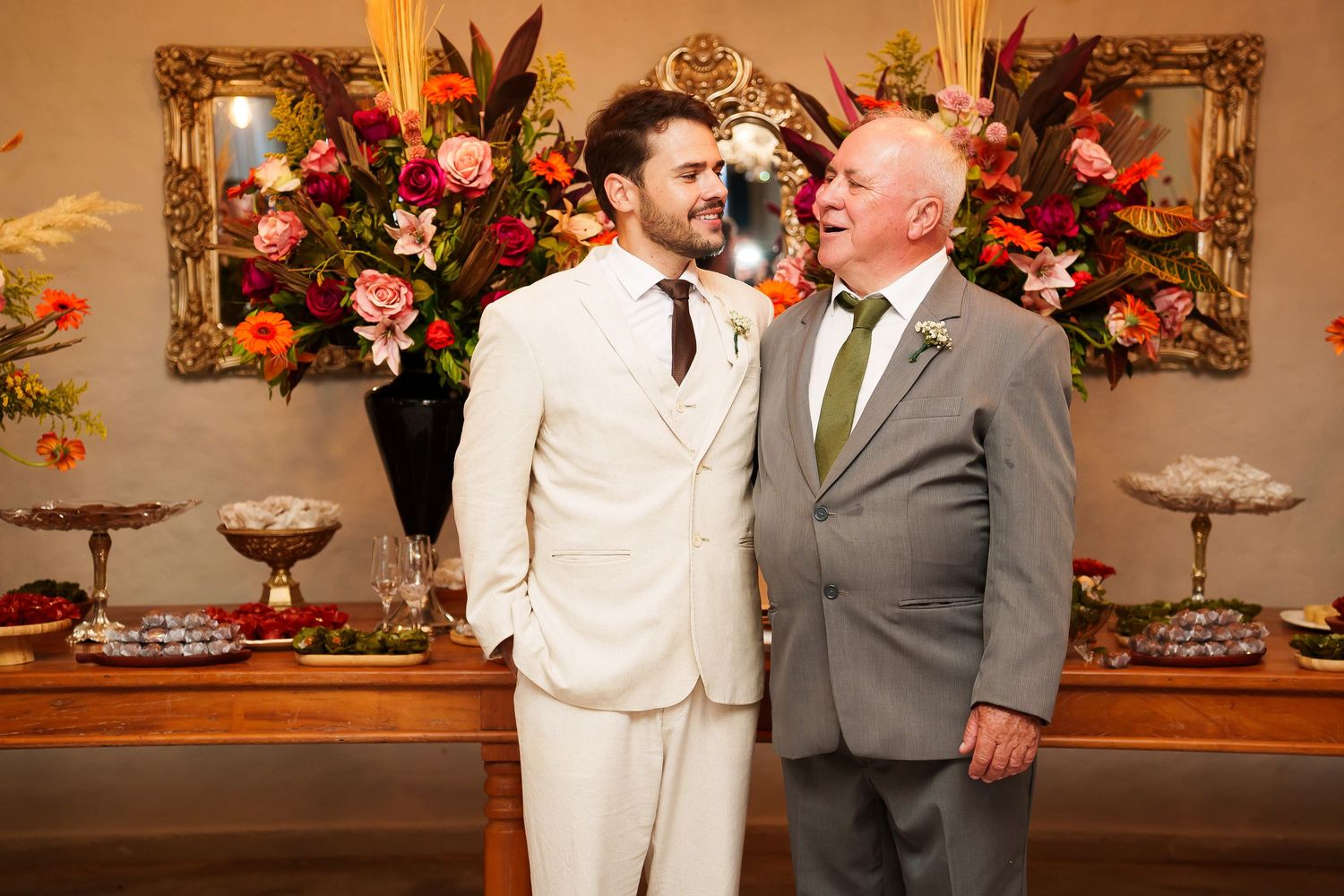 Two men in suits pose together at a wedding reception with colorful floral arrangements in the background.