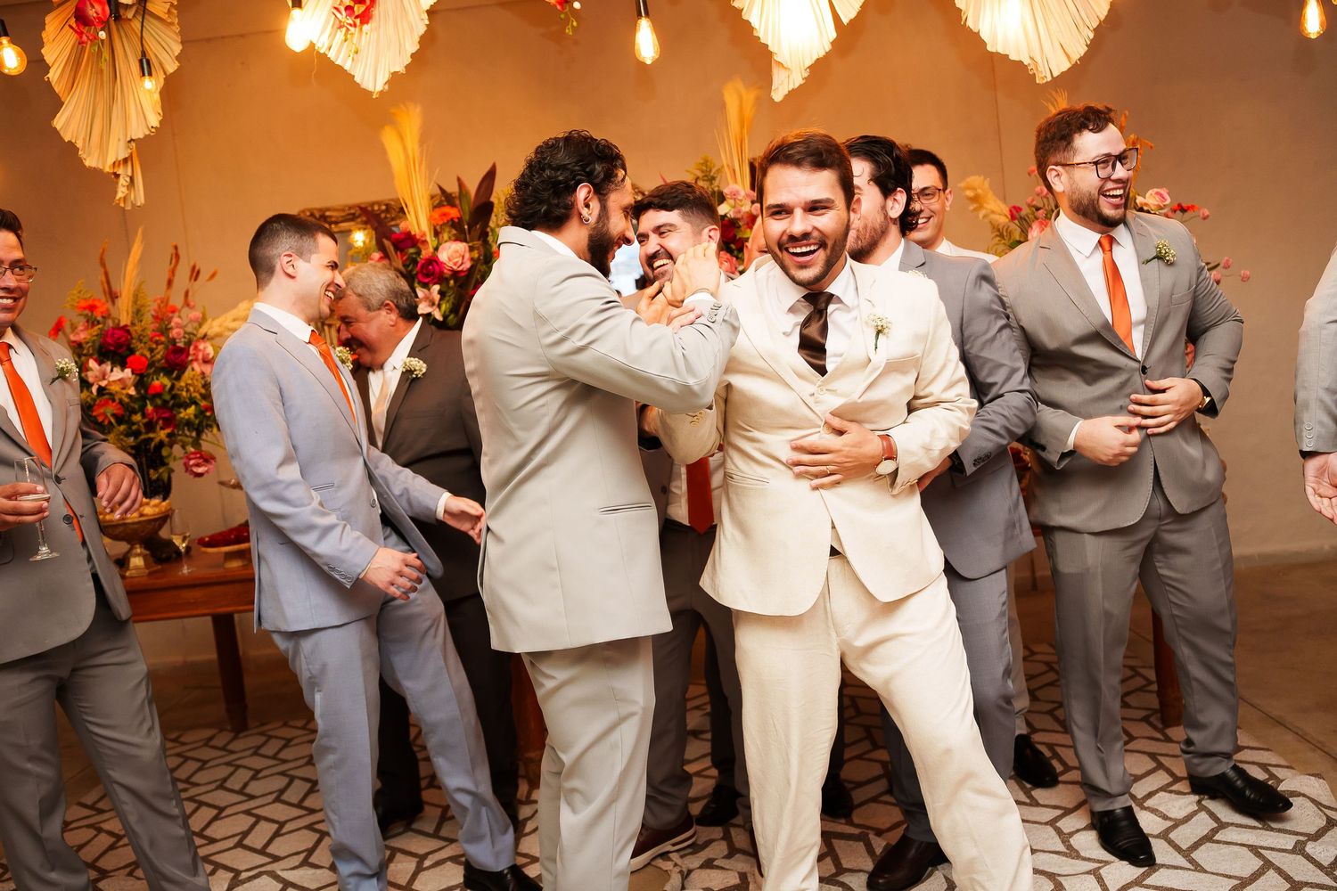 Groomsmen in gray suits celebrate with the groom in a cream suit at a colorful wedding reception.