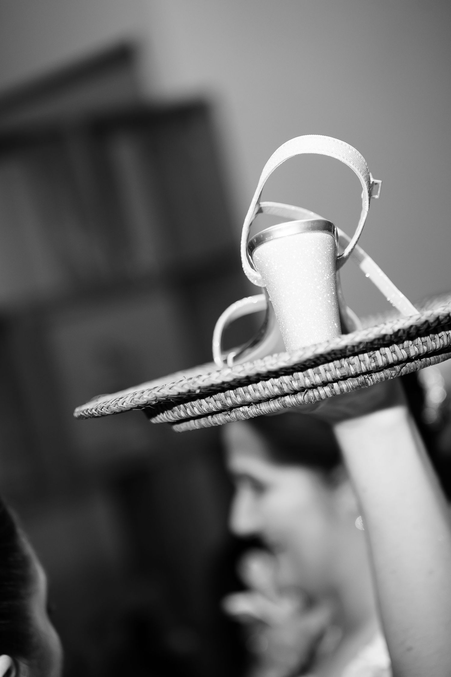 Black and white photo of a strappy sandal heel balanced on a straw hat, with a blurred person in the background.